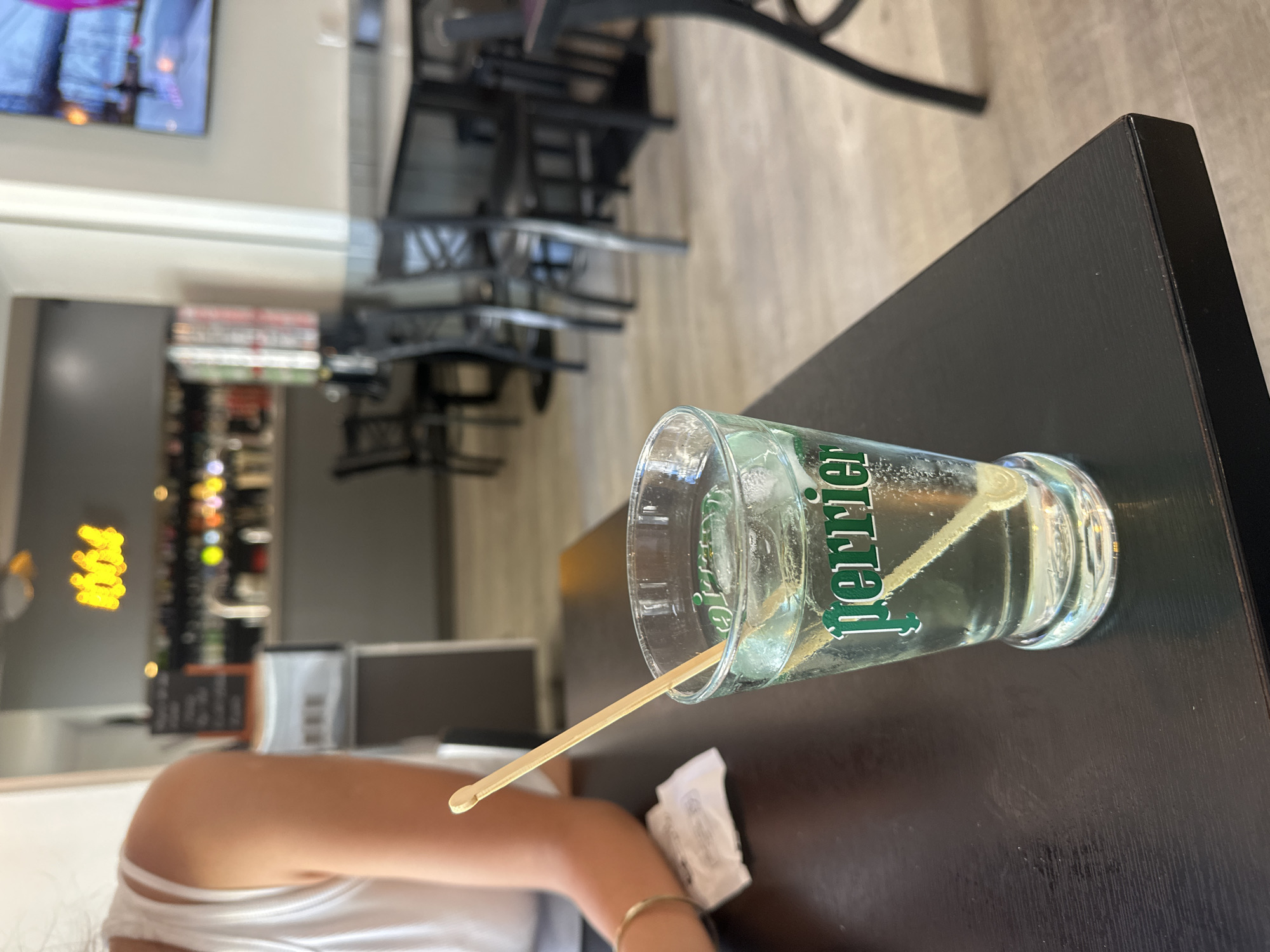 A glass of Perrier with ice and a wooden stirrer sits on a dark table in a restaurant. In the background, a woman wearing a white top is partially visible on the left. Further back, a bar area with bottles and a neon sign reading "1664" is visible. Chairs and tables are arranged in the dining area. The flooring appears to be light-colored wood.