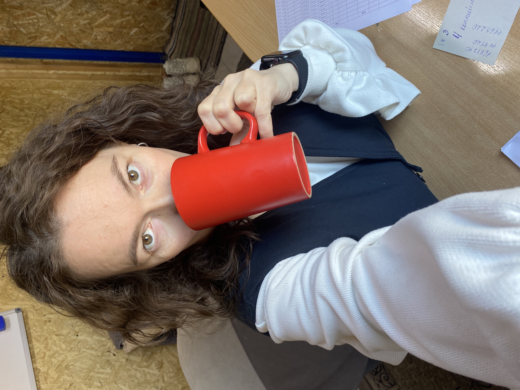A woman with curly brown hair is holding a red mug to her mouth. She is wearing a white long-sleeved shirt under a navy blue vest. A smartwatch is visible on her left wrist. The background includes a wooden desk with papers on it, a partially visible wall with what appears to be paneling, and a glimpse of a carpet. The location is Ol'khovskoye, Russia.