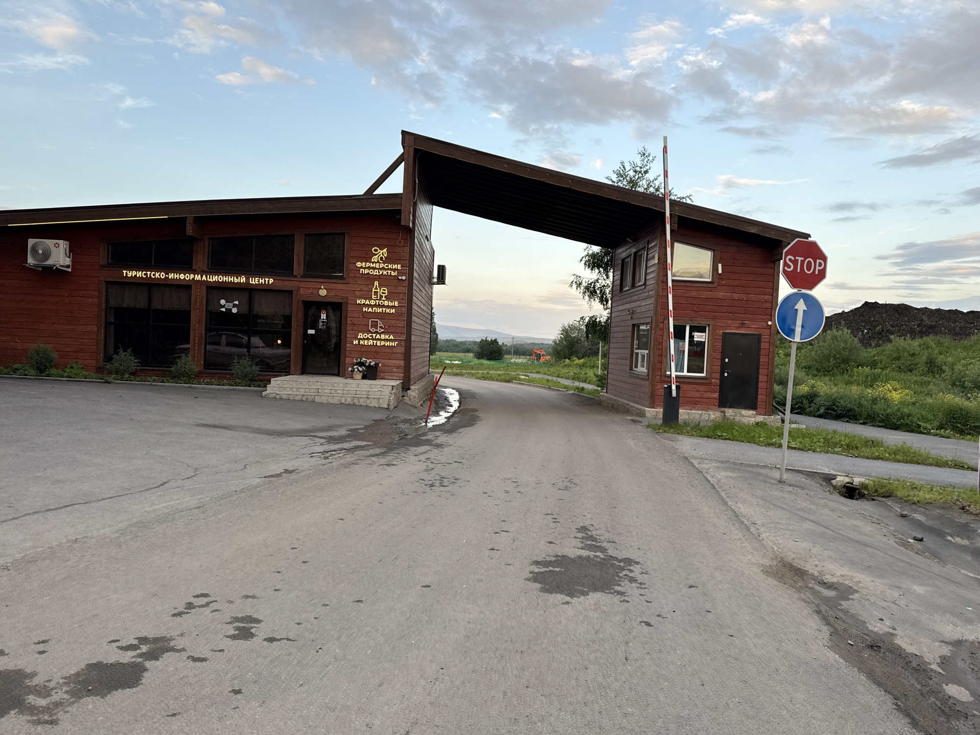 The image shows a wooden building with multiple windows and doors, an air conditioning unit mounted on the exterior, and signage indicating it is a tourist-information center. Signs on the building also advertise "farm products," "delivery," and "drinks." A red and white striped barrier arm is lowered across a paved road in front of the building. A blue circular sign with a white arrow pointing right and a red octagonal stop sign are visible on a pole near the barrier. The ground appears wet in places, with dark asphalt and patches of water or oil. The building is situated in a rural or semi-rural environment with green grass and trees visible. In the background, a construction vehicle can be seen. The sky is partly cloudy. This scene is located in the city of Syrostan, Russia.