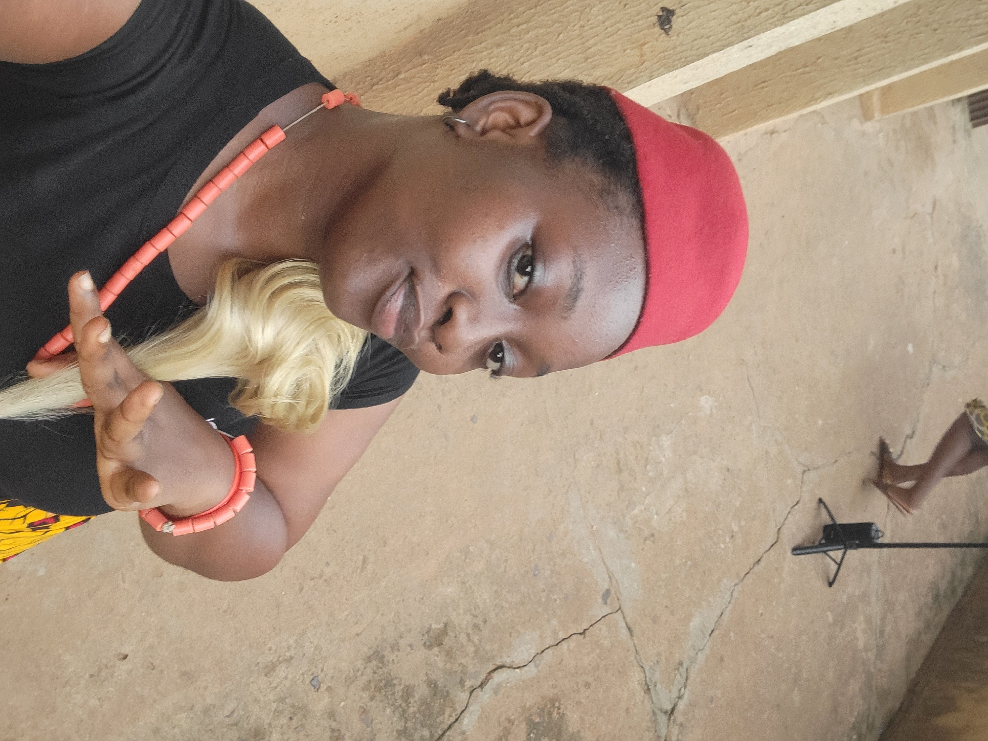 A woman wearing a black shirt, red cap, and coral beads around her neck and wrist, with a blonde wig, is indoors on a concrete floor. In the background, a tripod and bare feet are visible. The location appears to be in the city of Onitsha, Nigeria.