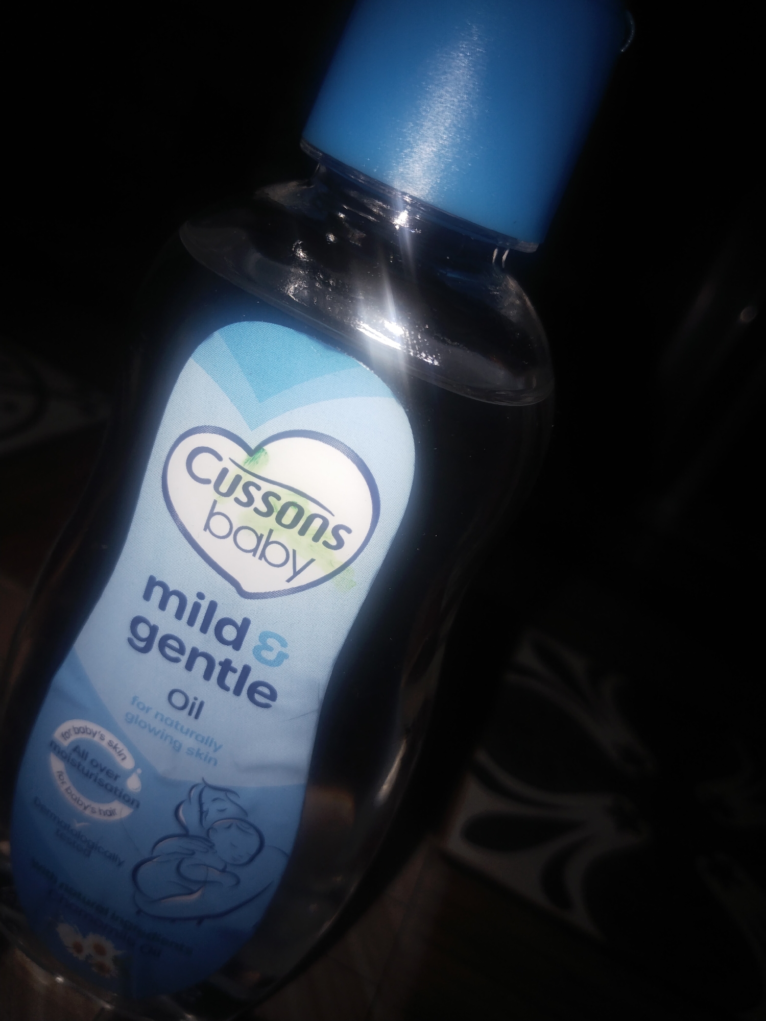 The image shows a bottle of Cussons Baby Mild & Gentle Oil. The bottle is clear plastic with a blue cap. The label is light blue with white text, featuring the brand name "Cussons Baby" in a heart shape. The product is described as "mild & gentle Oil for naturally glowing skin." A small image of a mother holding a baby is on the lower part of the label. The background is dark, suggesting an indoor setting. The oil is marketed for baby's skin and hair, claiming "all over moisturisation." The bottle appears to be on a dark surface. The image provides no details about the specific location in Uyo, Nigeria.