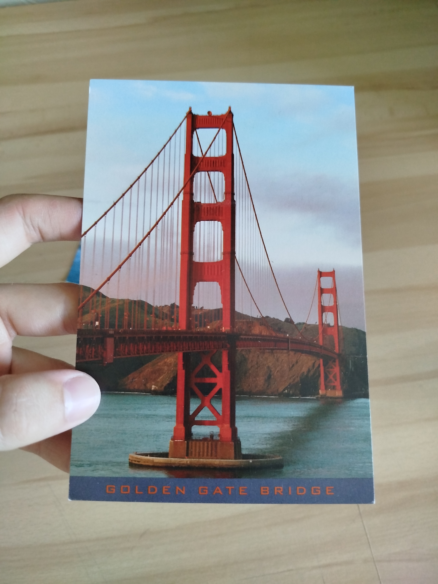 The image is a postcard featuring the Golden Gate Bridge. The bridge is depicted in a vibrant red hue against a backdrop of a partly cloudy sky and a body of water. Hills covered in vegetation are visible on either side of the bridge. The bottom of the postcard has text that reads "GOLDEN GATE BRIDGE". A person's hand is partially visible holding the postcard from the left side. The postcard is placed on a wooden surface. There is no indication that this scene is located in the city of Varna, Bulgaria.