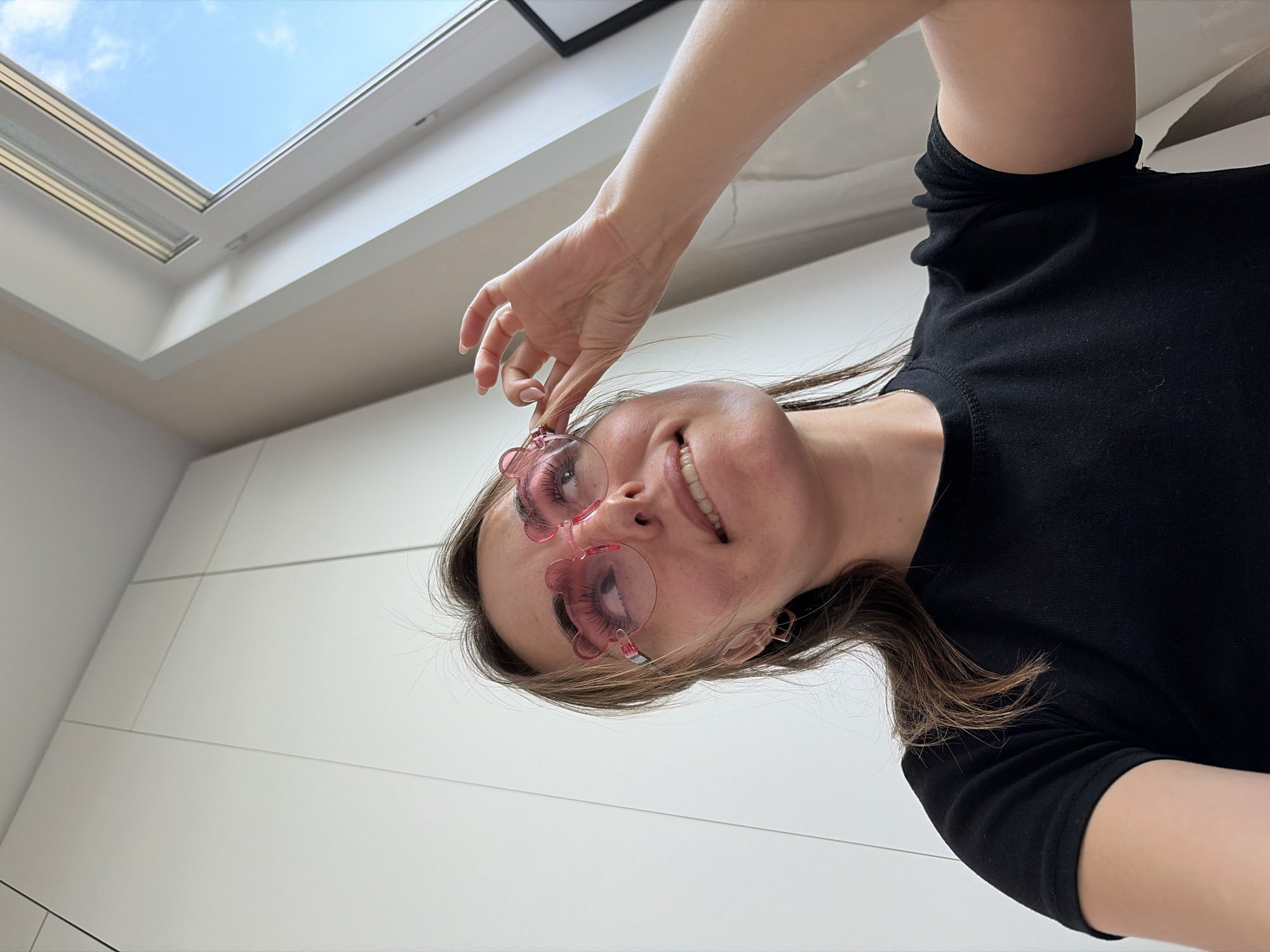A woman wearing pink flower-shaped sunglasses and a black T-shirt is holding the glasses with her right hand. She is smiling and appears to be indoors, judging by the white wall panels and skylight. The skylight shows a blue sky with some clouds. The location is not discernible from the image and therefore cannot be determined to be Izhevsk, Russia.