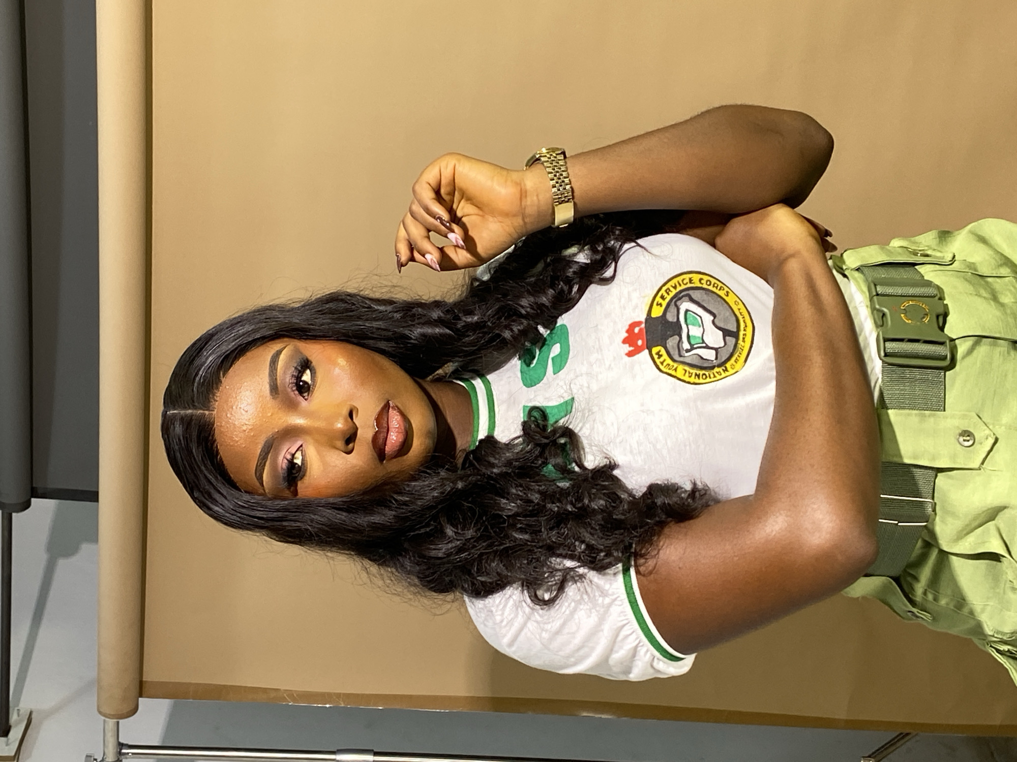 A young Black woman with long, dark, curly hair and full makeup is posing in front of a light brown backdrop. She is wearing a white t-shirt with green trim and a circular emblem on the front that appears to depict a map of Nigeria with a shield. She is also wearing olive green pants and a matching belt with a large buckle. Her arms are crossed, and she is looking off to her right with a thoughtful expression. A gold watch is visible on her left wrist. The lighting is soft, creating a warm glow on her skin. The background is plain and uncluttered, with a hint of photographic equipment on the left side of the frame. The setting appears to be a studio or an indoor photo shoot. There is no information in the image to indicate the location in Gura Top, Nigeria.