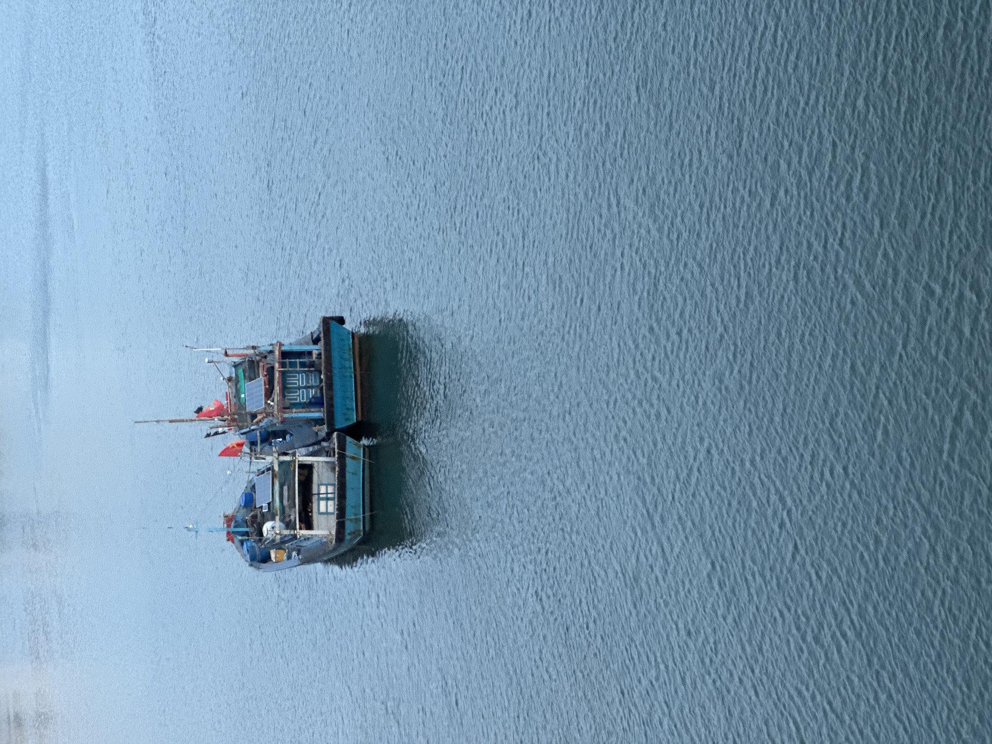 Two blue fishing boats are docked in the water. The boats are equipped with solar panels, masts with red flags, and small cabins. The water is calm with small ripples. The overall tone of the image is overcast and muted. This is likely in Đà Nẵng, Vietnam, as this type of fishing boat is common in the region. The exact location within Đà Nẵng cannot be determined from the image alone.