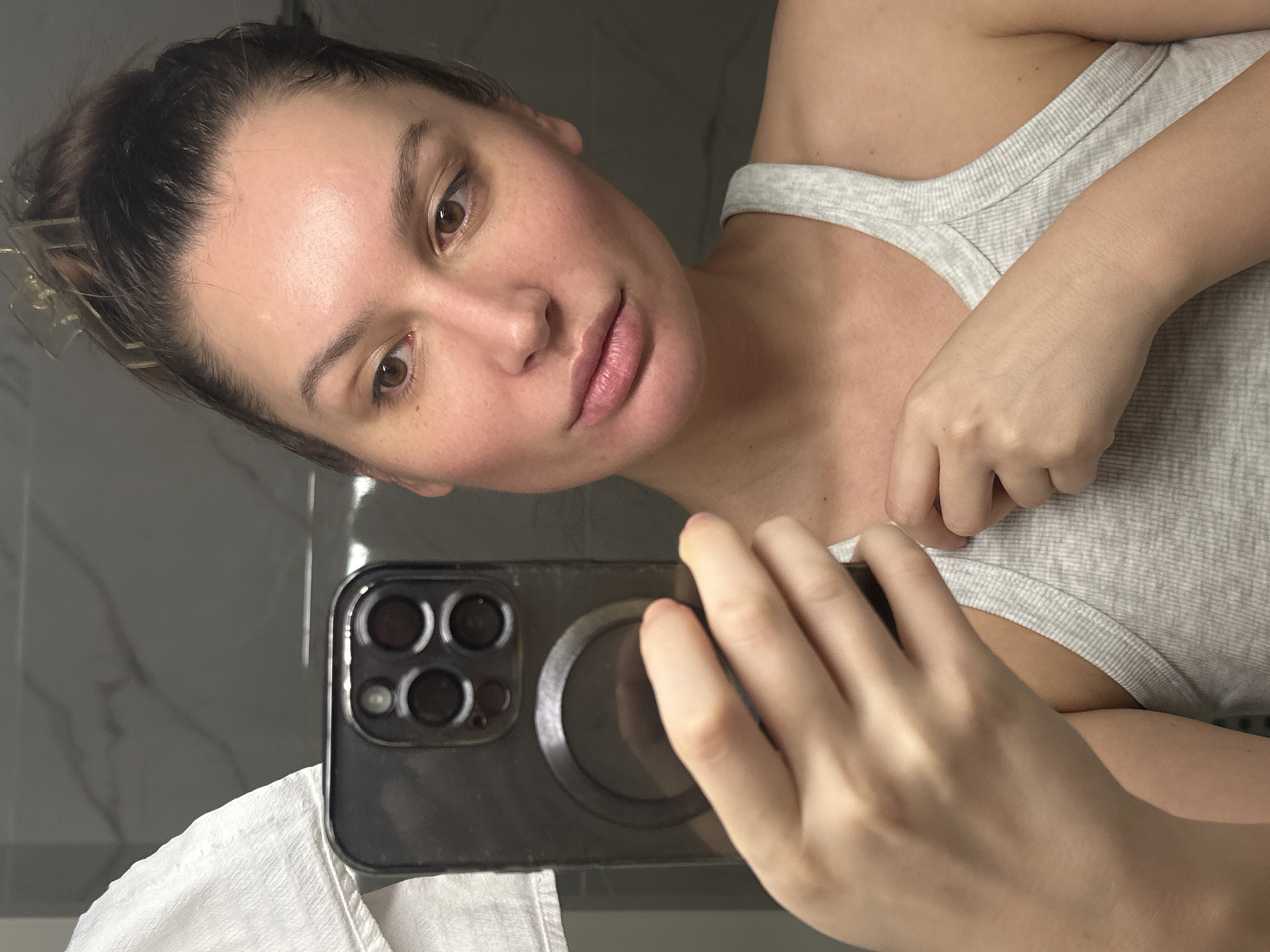 A young adult woman is captured in a mirror selfie, holding a dark-colored smartphone in her right hand. Her head and body are tilted, with her gaze directed towards the phone's camera reflecting her image. She has dark hair, brown eyes, and a light to medium skin tone with visible redness on her face, particularly on her cheeks and forehead. Her lips are full and naturally pink. She is wearing a light grey, ribbed tank top. Her left hand is subtly touching the strap of her top.
The smartphone features a distinct rear camera array and a circular magnetic accessory (likely MagSafe). In the reflected background, behind the woman, there is a smooth, dark grey surface with subtle white veining, possibly a marbled wall or counter. A white towel is partially visible in the bottom left corner of the frame. The lighting is bright and even, suggesting an indoor setting like a bathroom or vanity. The woman's expression is neutral and calm. No visible text is present in the image.