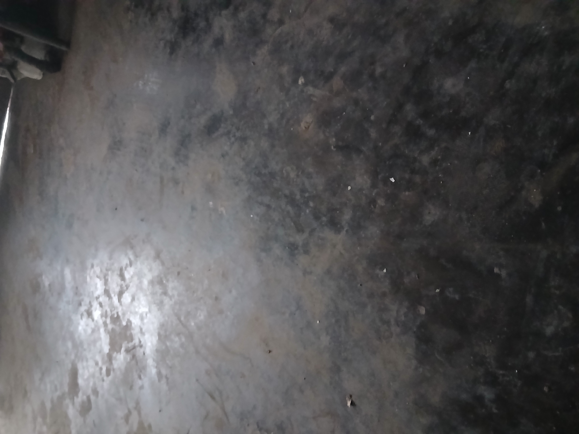 The image depicts a close-up, high-angle view of a dark, textured floor, likely concrete or asphalt, showing variations in tone and patches of lighter discoloration. The floor appears worn and possibly damp in some areas, with small debris scattered across its surface. The lighting creates a somewhat reflective sheen on parts of the floor, obscuring finer details. In the upper right corner, a blurred segment of what appears to be a metallic object, possibly a vehicle part or machinery, is visible. The overall environment seems industrial or utilitarian, with no discernible human presence or specific landmarks of Kademangan Kulon, Indonesia.