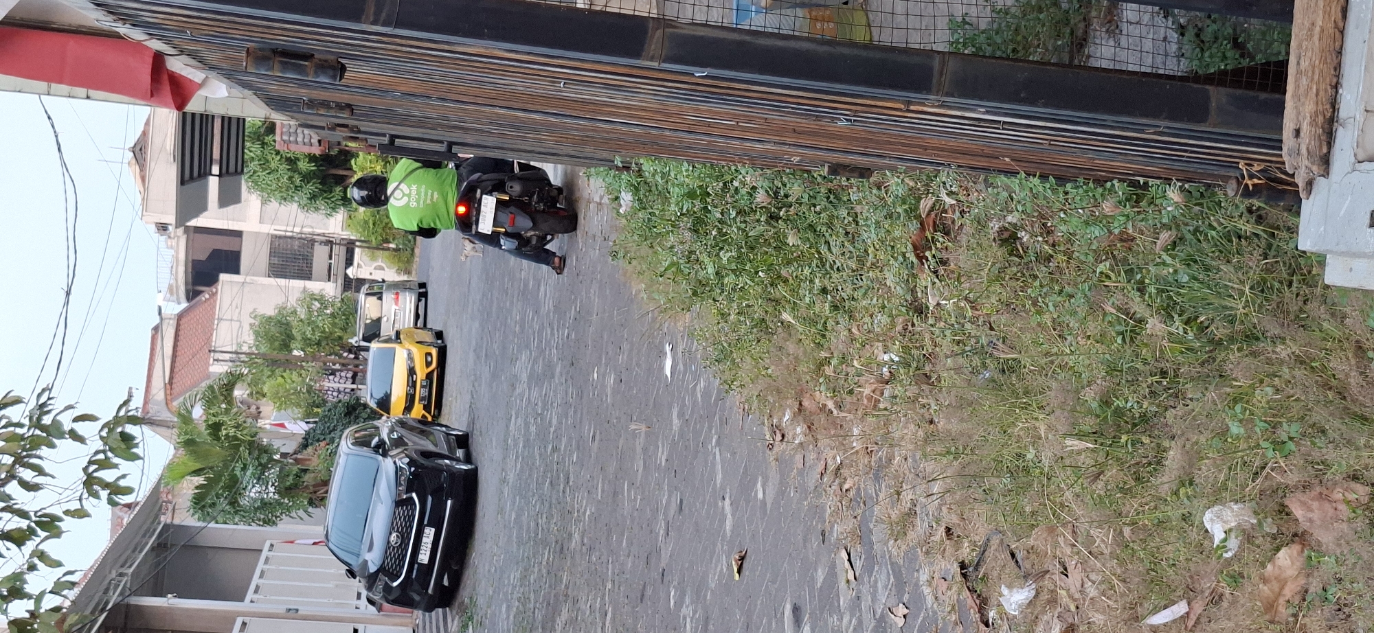 A motorbike rider wearing a green jacket with the Gojek logo is seen from behind, traveling down a paved alley in Surabaya, Indonesia. The rider is wearing a black helmet and blue jeans. A black SUV is parked on the left side of the alley, and a yellow car is driving ahead of the motorbike. Buildings with tiled roofs line the street, and lush green foliage grows along the sides of the alley, with some dry grass and fallen leaves on the ground. A metal fence runs along the right side of the image.