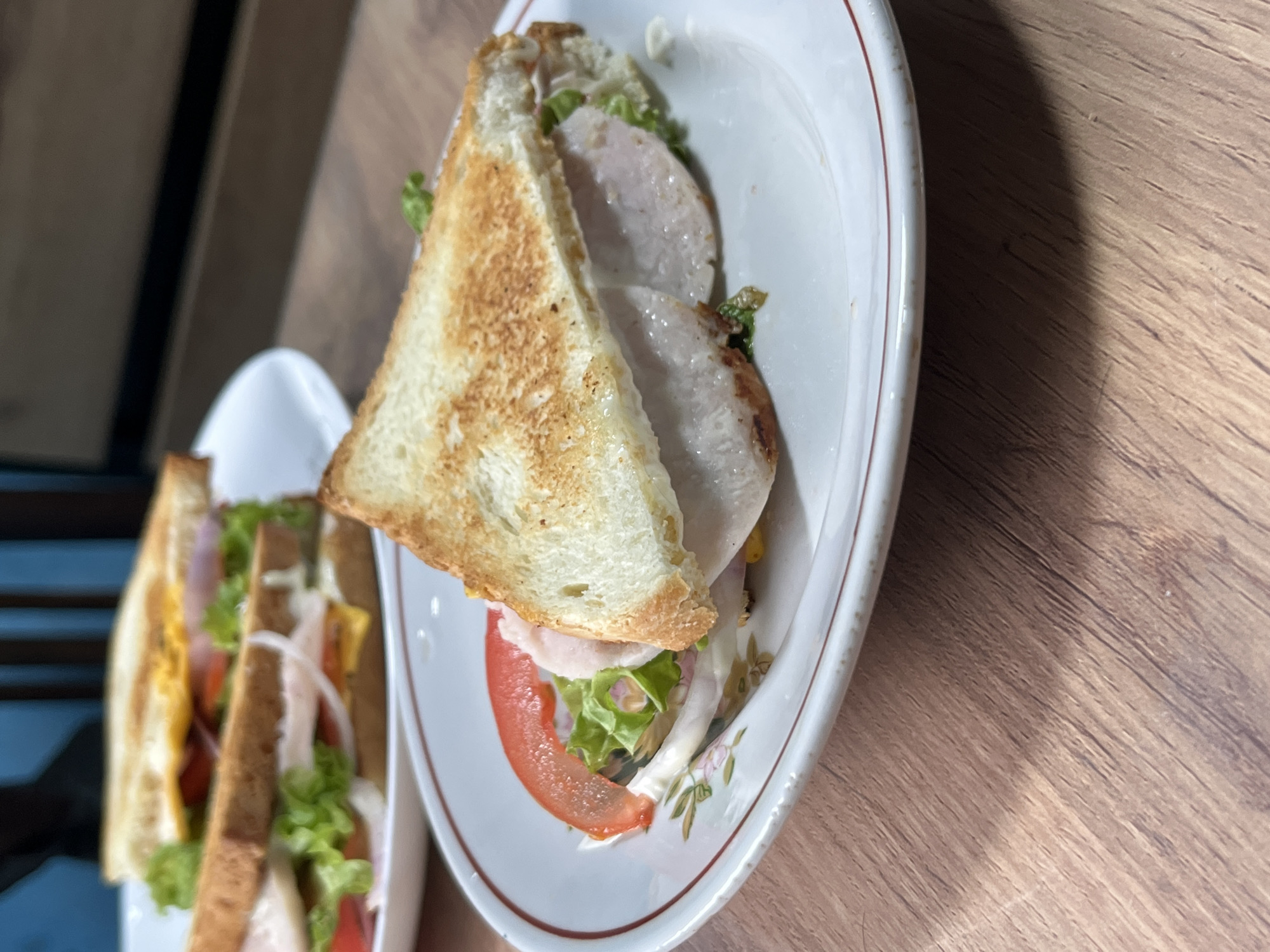 Two sandwiches are presented on white plates with a red rim. The sandwich in the foreground is sliced in half diagonally, revealing layers of toasted bread, lettuce, tomato, onion, and what appears to be turkey slices. The second sandwich, visible in the background and slightly out of focus, is also on a white plate and appears to be a similar club sandwich with similar ingredients. The sandwiches are arranged on a wooden surface, likely a table. The lighting suggests an indoor setting, possibly a home or cafe, with soft light illuminating the food. There are no people visible in the image. The image does not provide any information that would allow for its location to be specified as Nizhny Tagil, Russia, or any other city.