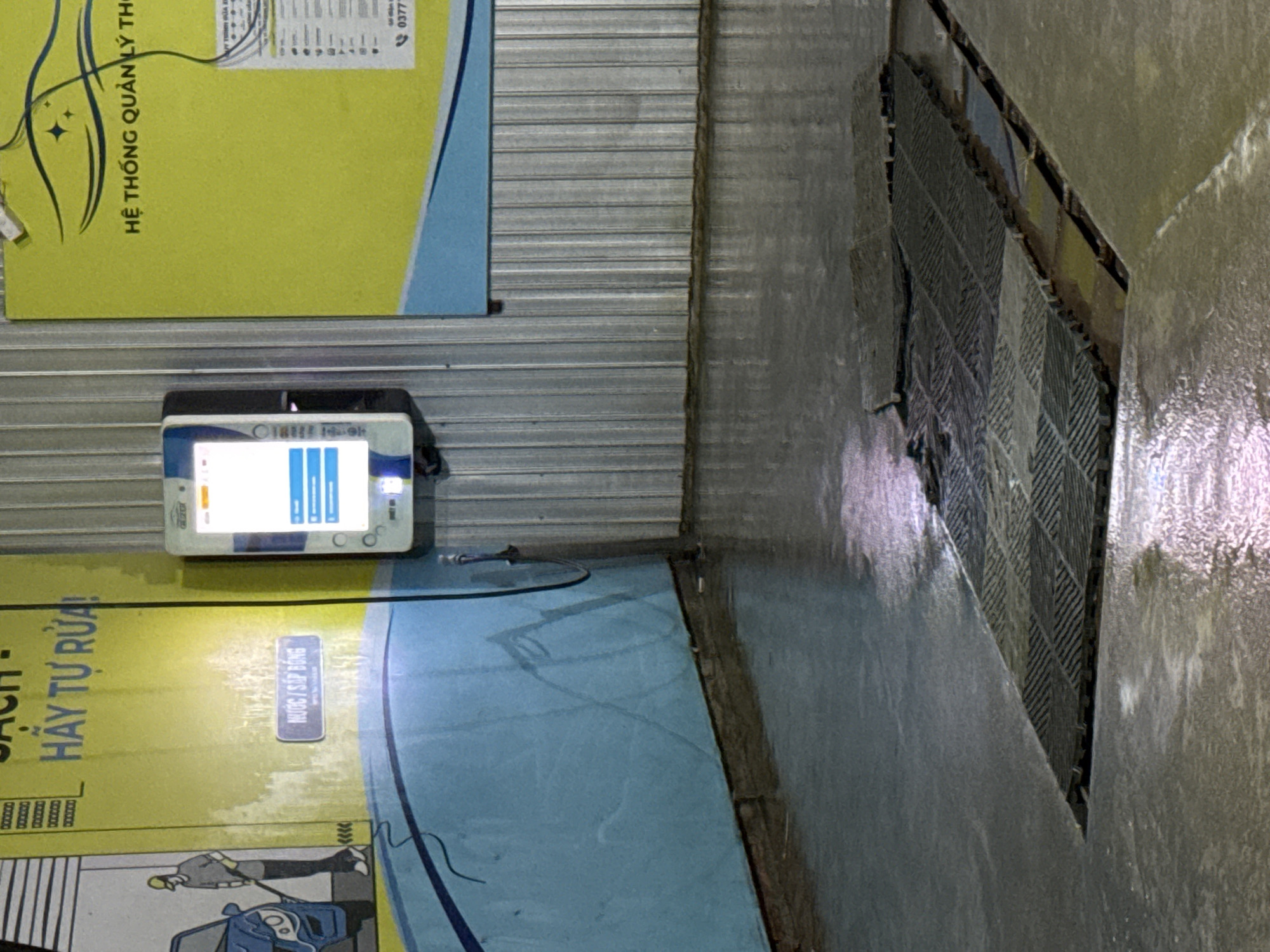 A self-service car wash area is depicted in the image, located in Bac Nha Trang Ward, Vietnam. The scene features a wet, tiled floor with visible puddles of water, indicating recent use. Several dark grey, interlocking rubber mats are laid out in the center of the car wash bay, presumably to provide grip. To the left, a faded yellow wall features a cartoon illustration of a person washing a blue car with a hose. Above the illustration, text in Vietnamese reads "HÃY TỰ RỬA!" (Wash it yourself!). A small blue button with Vietnamese text, possibly indicating a payment or service option, is also present on this wall.
To the right of the illustration, a white electronic payment or control terminal is mounted on a corrugated metal wall. The terminal's screen displays a bright white interface with some blue text and icons, suggesting it is operational. Further to the right, a large yellow sign with Vietnamese text, including "HỆ THỐNG QUẢN LÝ THU..." (Management System...), is visible, along with what appears to be a phone number, "0377..."
The environment is dimly lit, with reflections on the wet surfaces. The corrugated metal wall and the overall setup suggest a functional, utilitarian space designed for car washing. There are no people actively engaged in washing a car at the moment the photo was taken, but the presence of the equipment and the wet floor imply ongoing activity.