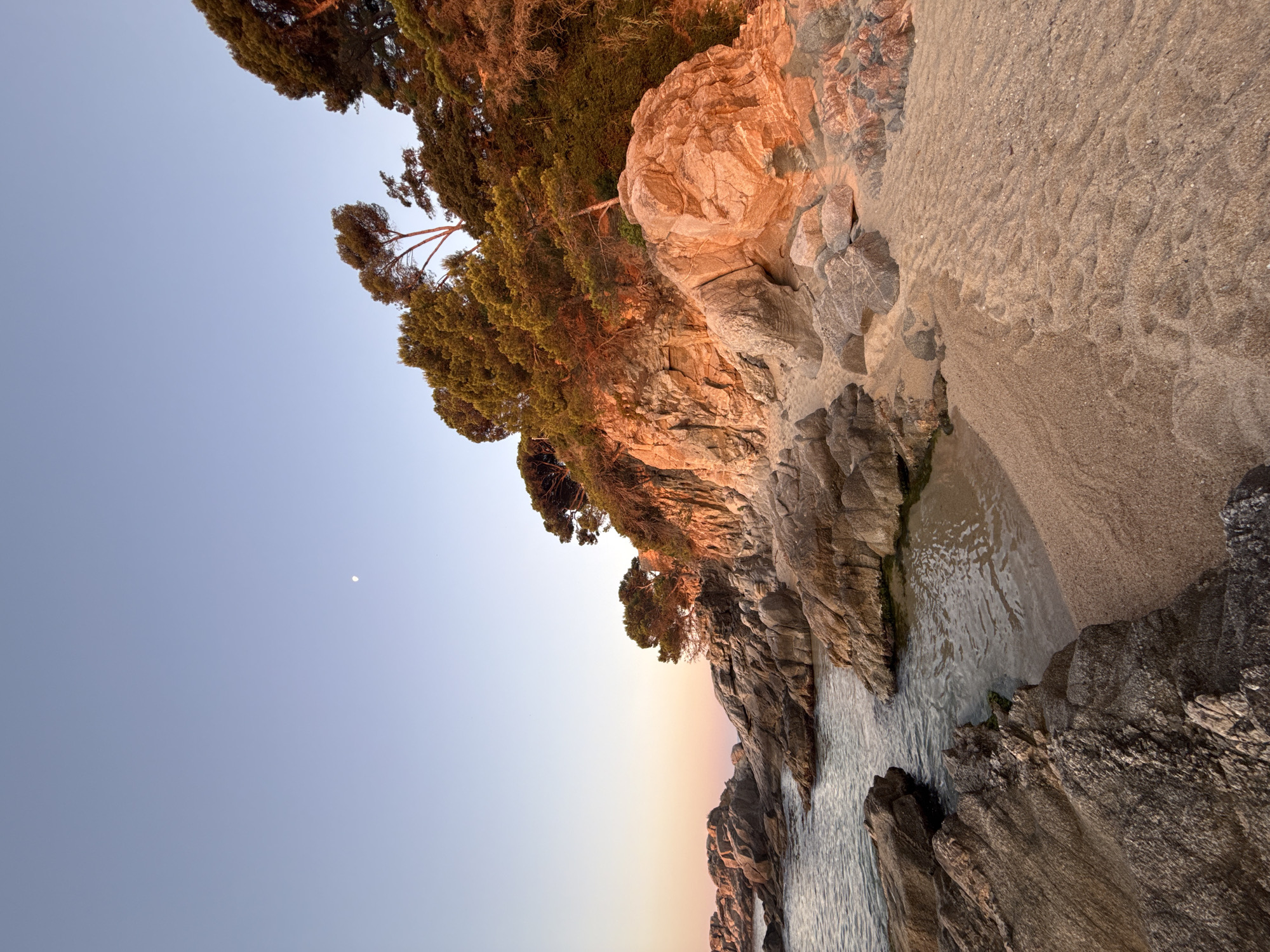A serene coastal scene unfolds at sunset in Castell d'Aro, Platja d'Aro i s'Agaró, Spain. A small crescent of sand, marked with subtle ripples, meets the gentle lapping of the sea against weathered, grey rocks. Beyond the beach, large, rugged rock formations rise, their surfaces bathed in the warm, golden hues of the setting sun. Lush green trees with dense canopies cling to the top of the cliffs, their foliage tinged with the same sunset glow. The sky above is a soft gradient of pale blue and delicate pink, with a faint, distant moon visible. No people or animals are present in this tranquil natural landscape.