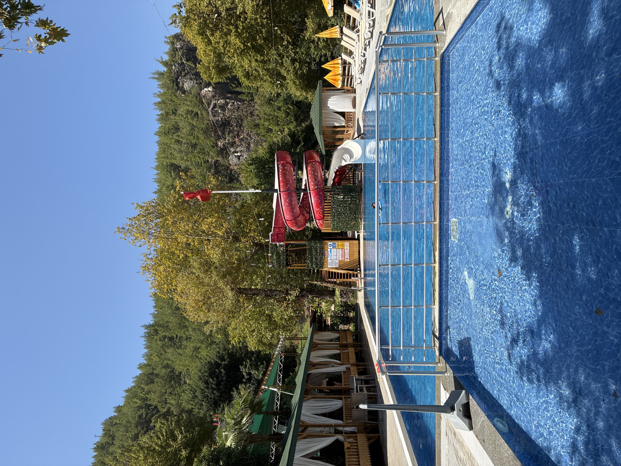 The image depicts a sunny day at an outdoor swimming pool complex located in Alanya, Turkey. In the foreground, a large, clear blue swimming pool fills the lower half of the frame, its tiled bottom visible through the shimmering water. A metal railing separates the foreground pool from a slightly deeper section of the pool in the background.
To the left of the pool, a covered seating area with white curtains offers shade. Further back, a vibrant red water slide spirals down from a wooden platform, ending in a white slide chute. Behind the water slide, a tall wooden structure with stairs and caution signs is visible. The background is dominated by a lush, green forested hillside under a clear blue sky. Yellow umbrellas and lounge chairs are arranged on the poolside, suggesting a recreational area.
A few people are visible in the water; one person is swimming in the background pool, and another appears to be descending the white slide. The overall scene conveys a sense of leisure and summer recreation.