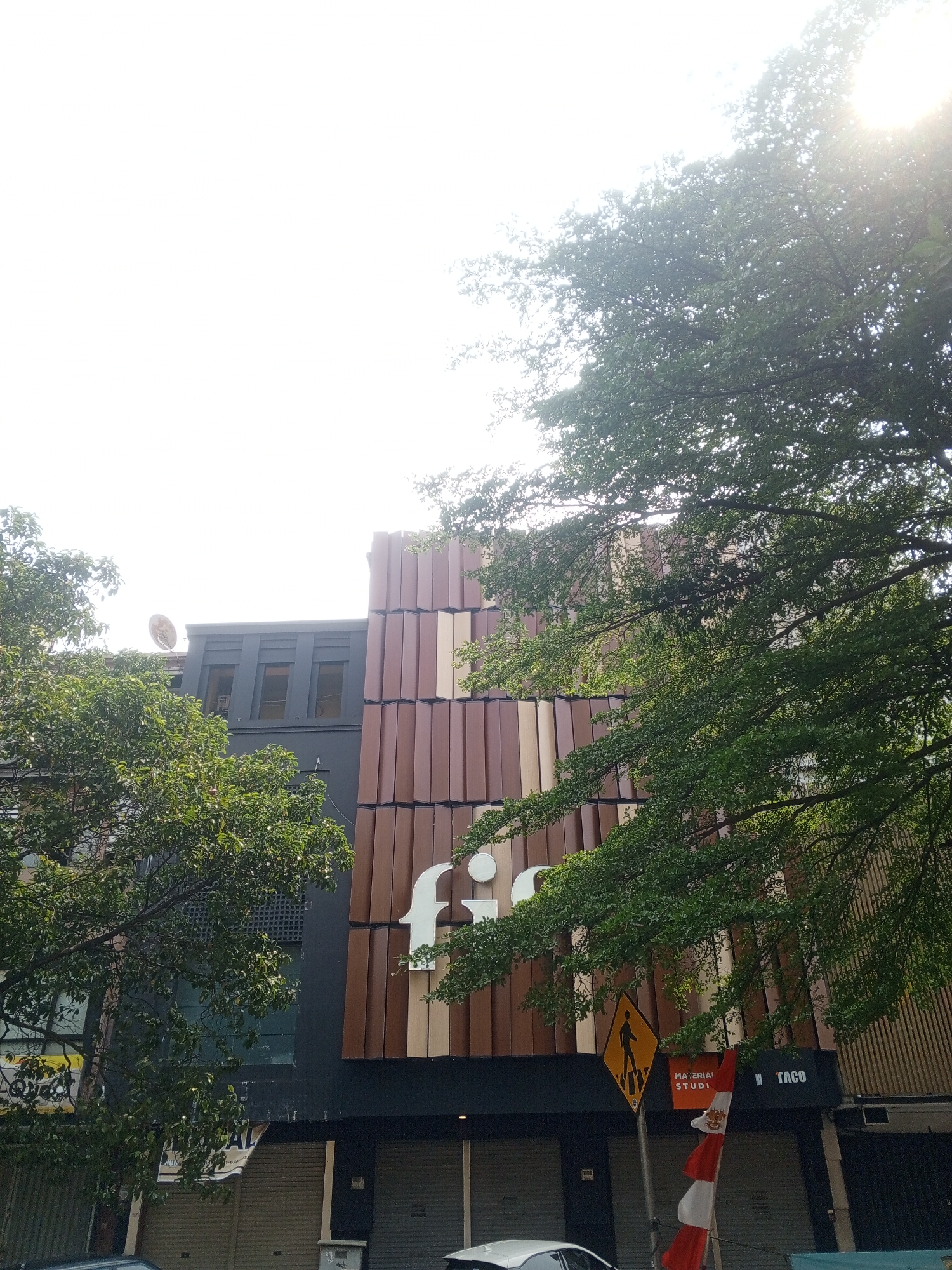 A low-angle exterior shot captures a building with a modern facade in South Jakarta, Indonesia, on a bright, sunny day. The building features a prominent sign with a white 'fi' logo against a backdrop of vertical brown and cream colored panels. To the left of the building, green trees with lush foliage partially obscure the view, with one branch extending into the foreground. A satellite dish is visible on the roof of an adjacent building, and a street sign with a pedestrian crossing symbol stands on the sidewalk. The ground level of the building has closed metal shutters. A red and white Indonesian flag is displayed on a pole. The sky is bright and washed out with sunlight.