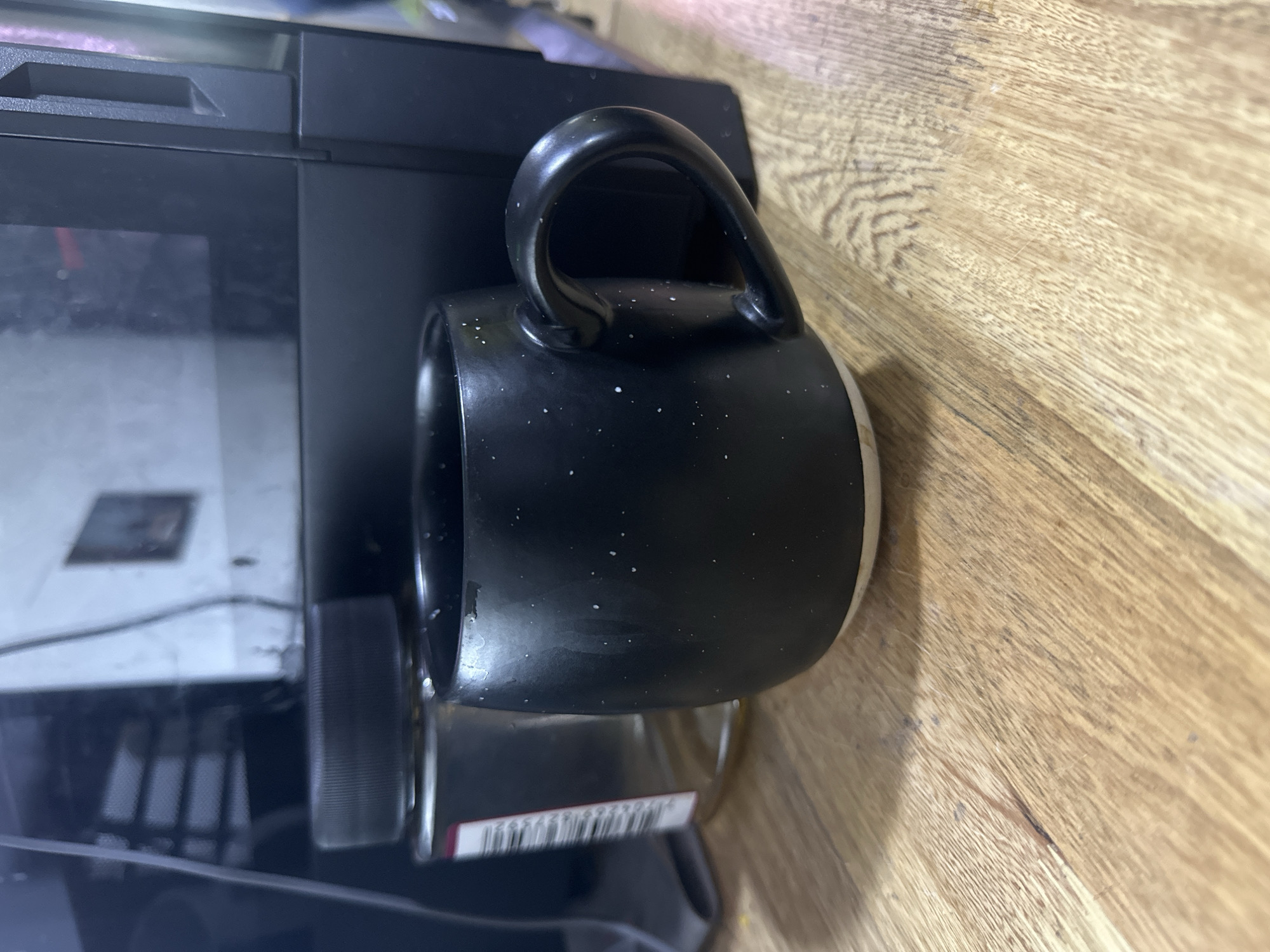 This is a close-up, rotated, eye-level shot of a black speckled mug resting on a light brown wooden surface. The mug has a curved handle and its interior appears to be lighter in color. Some white speckles are visible on the black exterior of the mug. Partially visible to the left of the mug is a black electronic device, possibly a printer or a computer tower, with a reflective surface. A barcode is faintly visible on a white label attached to a silver object beneath the mug. The wooden surface shows grain patterns and some slight discoloration or wear. The lighting appears to be artificial and casts subtle shadows. No people or animals are present in the image, and the scene is indoors. The overall mood is neutral, focusing on the objects and their immediate surroundings. No specific time of day or weather conditions are discernible. The location context "Juan Mina, Colombia" does not provide any visual cues within the image itself.
