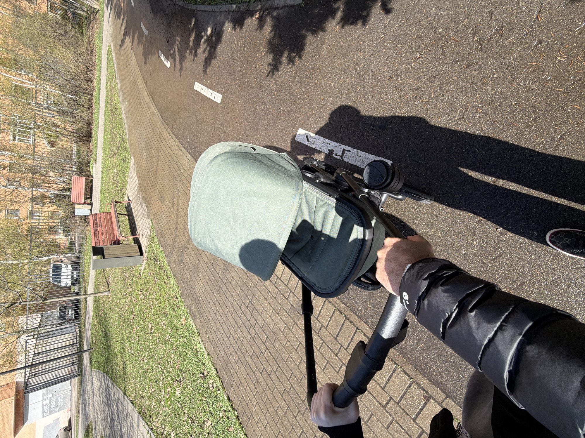 The image, taken from a first-person perspective, shows a person pushing a light green baby stroller along a paved path on a sunny day. The person's hands are visible on the stroller's black handle, and they are wearing a black puffer jacket with a small white emblem on the sleeve. Part of a dark, patterned shoe is also visible.
The paved path, made of light brown brick-like pavers, runs parallel to a strip of green grass. Adjacent to the paved path is a dark asphalt road or dedicated path, marked with white dashed lines. Strong shadows from overhead trees are cast across the asphalt, indicating bright sunlight. Scattered dry leaves or pine needles are visible on the ground.
In the background, behind the grass, are numerous trees and bushes, mostly bare of leaves, suggesting either early spring or late autumn. Further back, multi-story light-colored brick buildings with windows are visible, hinting at a residential area. A black metal fence runs along the property line, and a white car is partially visible behind it. Two red wooden benches and a grey trash can are positioned along the grassy area.
The time of day appears to be morning or late afternoon, given the length and angle of the shadows. The weather is clear and sunny. A small white triangular symbol is present on the stroller's adjustable handle. In the far background, some blurry Cyrillic text is discernable on a building or sign, which aligns with the provided context of Moscow, Russia. The overall scene depicts a calm, everyday outdoor activity in an urban or suburban setting.