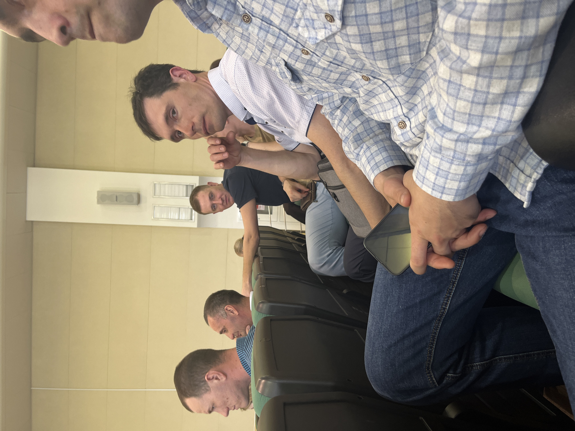 The image depicts several men sitting in rows of dark-colored chairs, possibly in an auditorium or lecture hall. The walls are a light tan color. One man is looking directly at the camera. A person is holding a phone. The location in Izhevsk, Russia cannot be determined from this image.