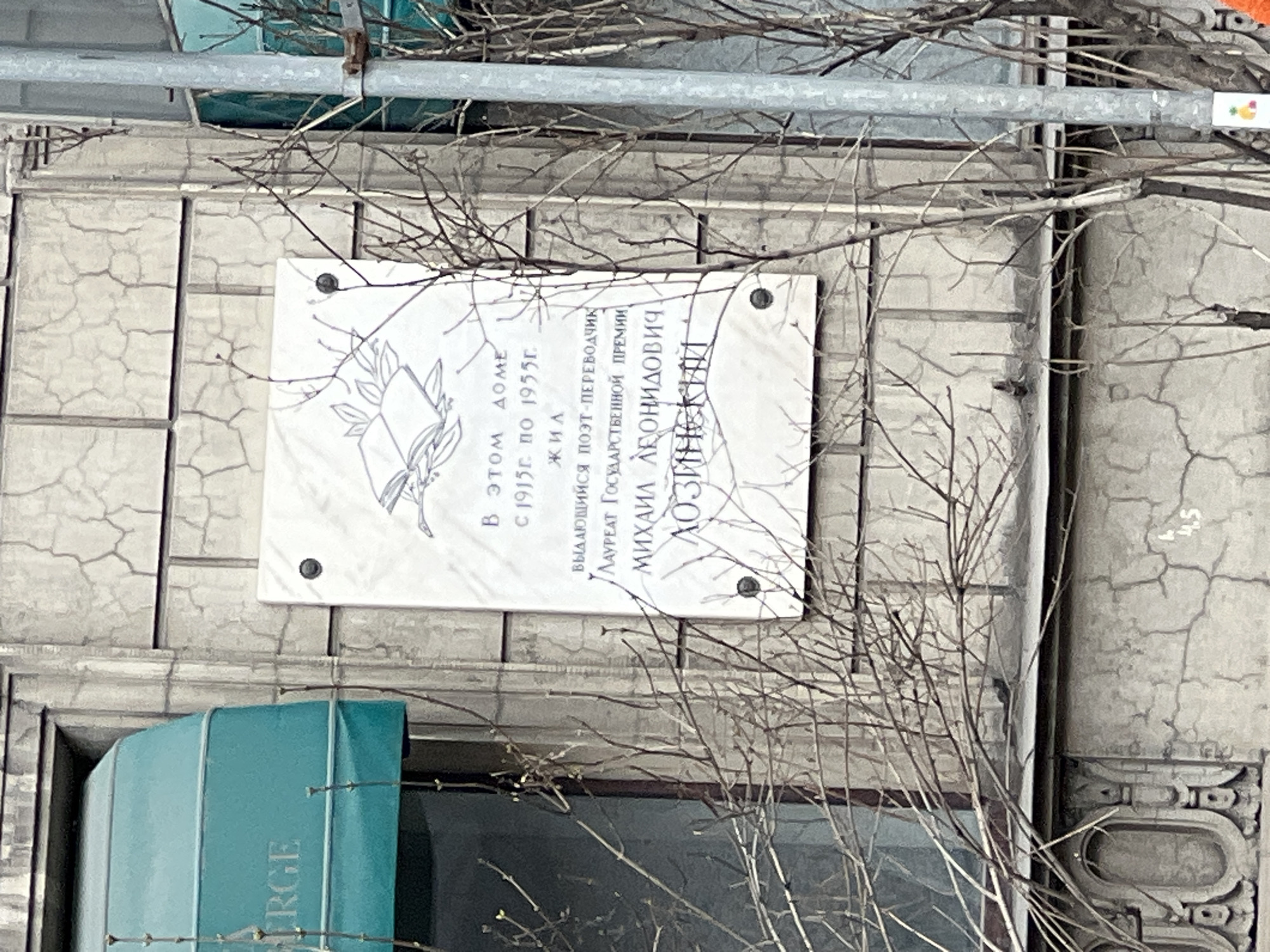 The image shows a plaque on a building, likely in Saint Petersburg, Russia. The plaque is dedicated to the poet and translator Mikhail Leonidovich Lozinsky, who lived in the building from 1915 to 1955. The plaque also mentions that he was a laureate of the State Prize. The plaque is mounted on a gray, weathered wall, with some branches obscuring part of it. The word "ERGE" is visible on a turquoise element below the plaque.