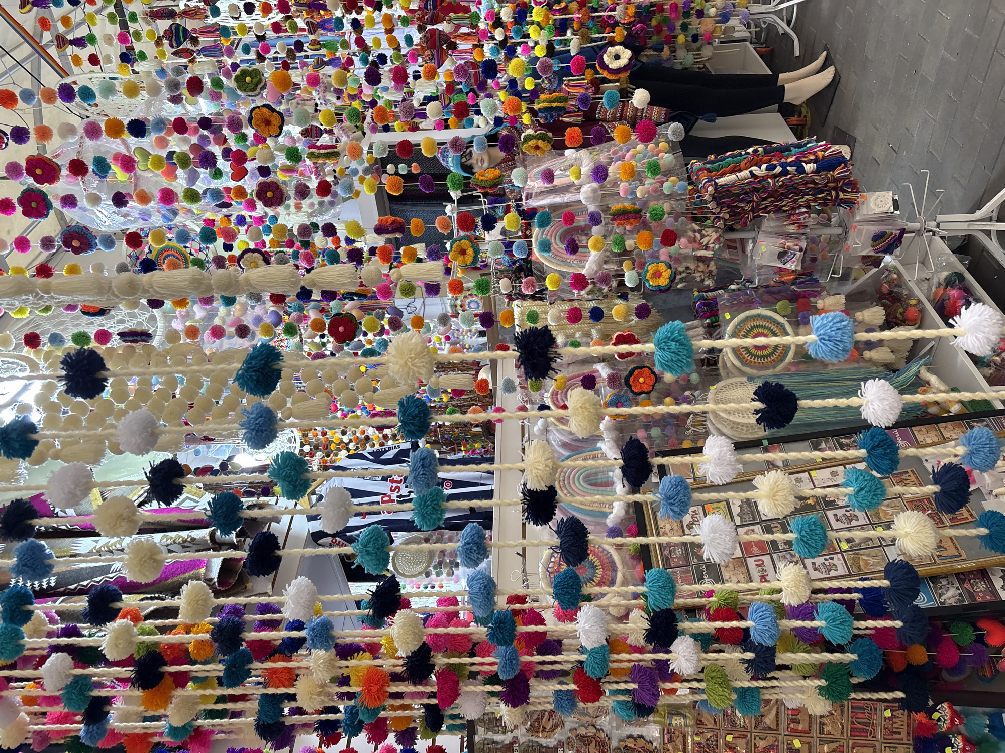 A stall in Miraflores, Peru, is filled with a vibrant and colorful display of handmade pom-pom and tassel decorations hanging from the ceiling and from display racks. The decorations are in a multitude of bright colors including red, orange, yellow, green, blue, purple, pink, and white, and are made of yarn. Some of the decorations are small pom-poms, while others are larger pom-poms or feature floral designs and tassels. In the background, a mannequin wearing a blue knit hat and a patterned scarf is visible, displayed amidst the hanging decorations. The ground appears to be paved with dark gray cobblestones.