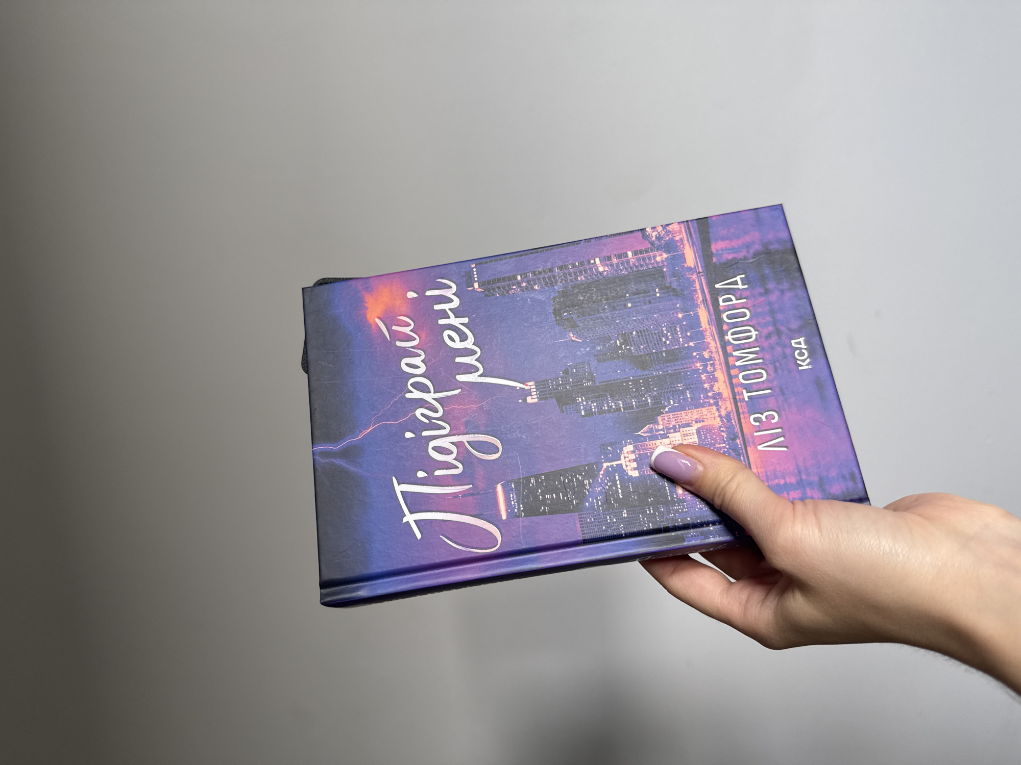 The image shows a person's hand holding a book. The book cover features a nighttime cityscape with purple and pink hues, suggesting twilight or evening. Lightning strikes are depicted in the background. The title of the book, written in a cursive font, is "Tigray i guerri" which translates to "Tigray and guerrillas". Below that, "ЛІЗ ТОМФОРД" and "КД" are visible, indicating the author or a publisher. The person's hand has a light skin tone, and their fingernails are painted with a light purple-white gradient. The background is a plain, light gray, likely a wall or backdrop. There are no strong indicators of weather conditions beyond the depiction of lightning on the book cover. The setting is not specified in the image itself, but external context indicates it is in Łódź, Poland.