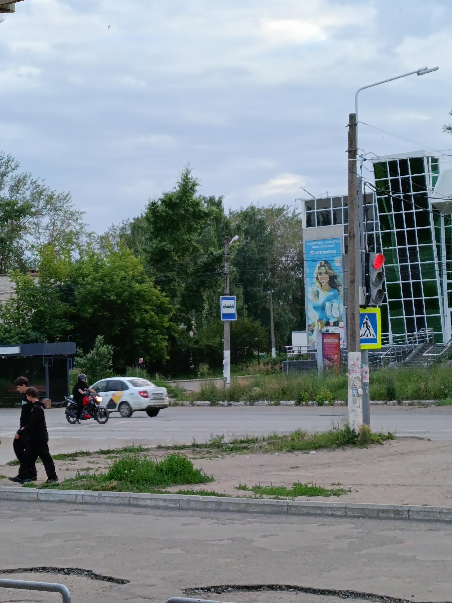 The image depicts a street scene in Chaikovsky, Russia, under an overcast sky. In the foreground, two young men in dark clothing walk side-by-side on a paved sidewalk. To their right, on the road, a person wearing a helmet rides a red and black motorcycle. A white sedan is driving in the same direction. Further in the background, a modern building with a distinctive grid-like glass facade stands tall. Adjacent to the building, a traffic light shows a red signal, and a pedestrian crossing sign is visible. A bus stop sign is also present. Green trees and vegetation are scattered throughout the scene, suggesting a suburban or semi-urban environment. A large advertisement featuring a smiling woman is displayed on the side of the building.