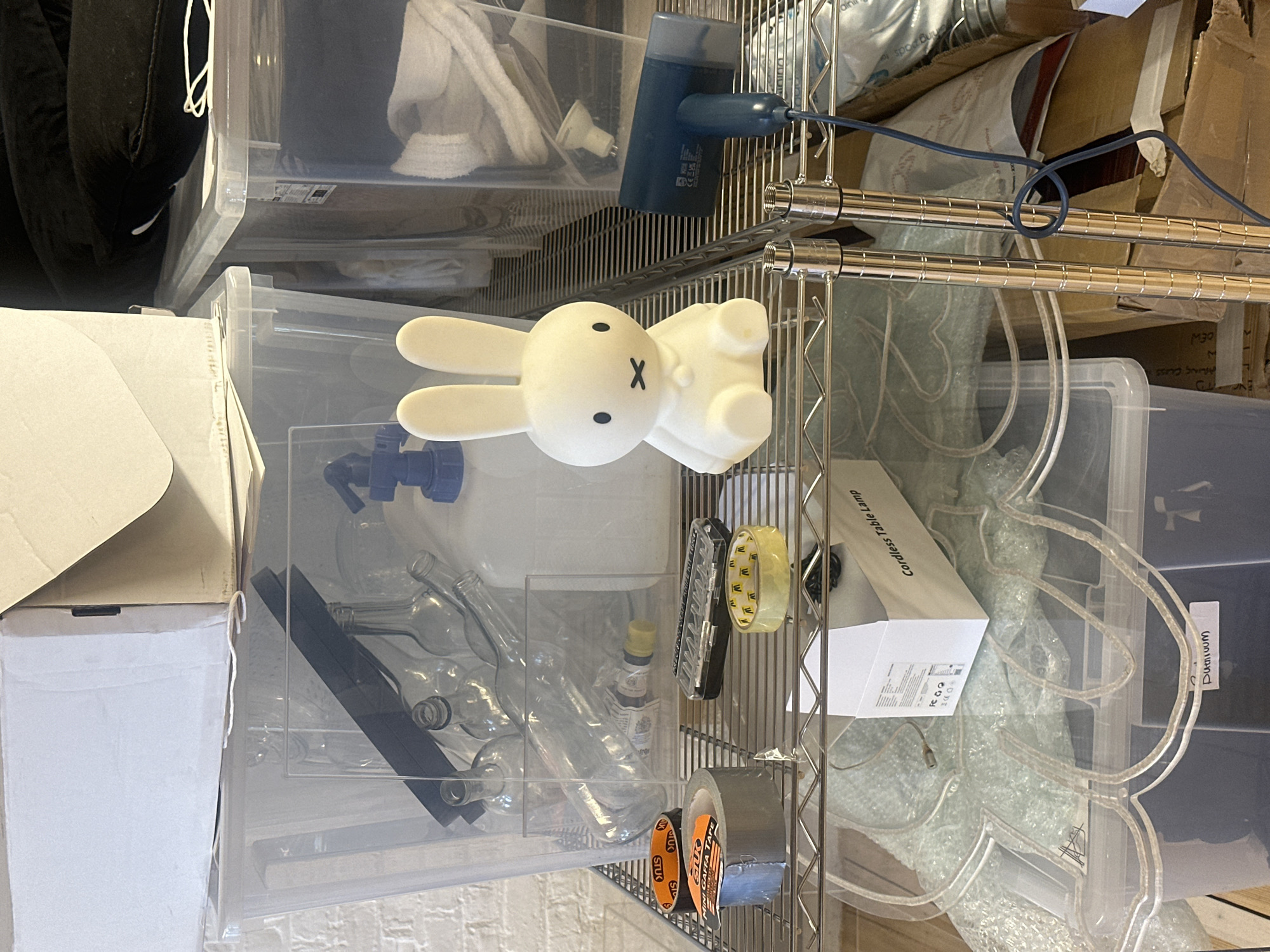 This image captures a close-up view of a metal wire shelving unit used for storage, filled with various items and containers. The overall scene suggests a cluttered storage area, possibly in a garage, utility room, or basement.
In the center-left, a prominent white Miffy bunny-shaped lamp rests on a clear plastic storage bin. This bin contains several empty glass bottles, some with spigots, and a small bottle with a yellow liquid. Above this, another clear bin holds black fabric and a white towel. To the right of the Miffy lamp, a blue device, possibly a hair dryer or an air pump, is plugged into a wall adapter and hangs from the shelving unit.
The shelves themselves are made of polished chrome or silver-colored metal. Below the Miffy lamp, on a lower shelf, a roll of gray duct tape and two rolls of orange tape labeled "STUK GAFFA TAPE" are visible. Next to these is a clear tape dispenser with yellow tape and a black case.
On the bottom right, another large clear storage bin holds bubble wrap, a white box labeled "Cordless Table Lamp," and what appears to be a clear, illuminated outline of a figure or design, possibly a neon sign. A label on this bin reads "Dunivum." In the background, various cardboard boxes and packaging materials are stacked. A black cable is coiled and draped over the shelving unit in the upper right.
No people are visible, and no specific activity is taking place; the image is a still life of stored household or workshop items.