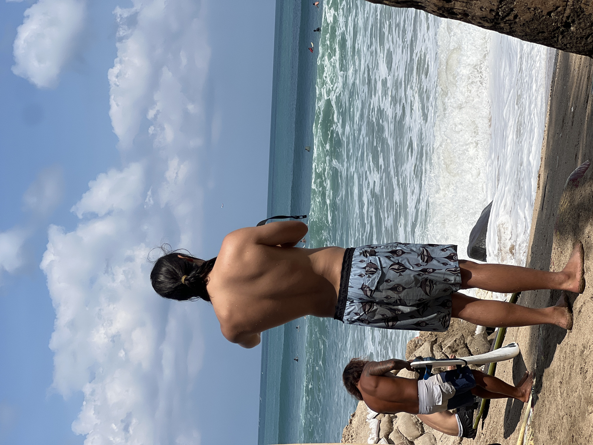 The image depicts a sunny day at Kuta Beach in Bali, Indonesia, an area known for its popular surf breaks. In the foreground, two individuals are present. The primary subject, a person with long dark hair tied back, stands facing the ocean, wearing patterned swim trunks and holding what appears to be a camera strap. To their left and slightly behind, another individual, shirtless and with visible tattoos on their back, is crouched down, holding a surfboard upright. The ocean is visible in the background with several surfers on the water, riding moderate waves. The beach itself is sandy, with some rocks and vegetation visible along the shoreline. The sky is a clear blue with scattered white clouds, and a small airplane is faintly visible in the distance. The overall scene suggests a casual beach day with recreational activities.