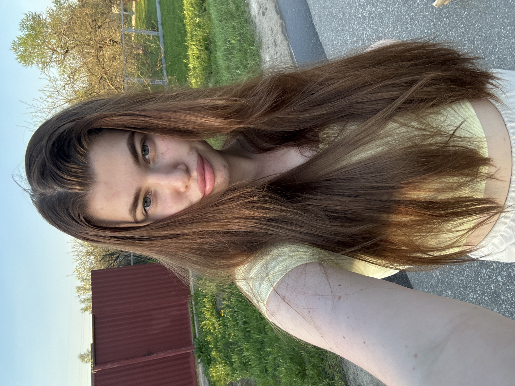 A young woman with long brown hair, wearing a yellow top, is taking a selfie outdoors. The background includes green grass, a red fence, a tree, and what appears to be a paved road. The location is likely Шевлягино, Russia. The woman is smiling slightly. Her hair partially covers her face. Her left arm is visible. The lighting suggests it is daytime.