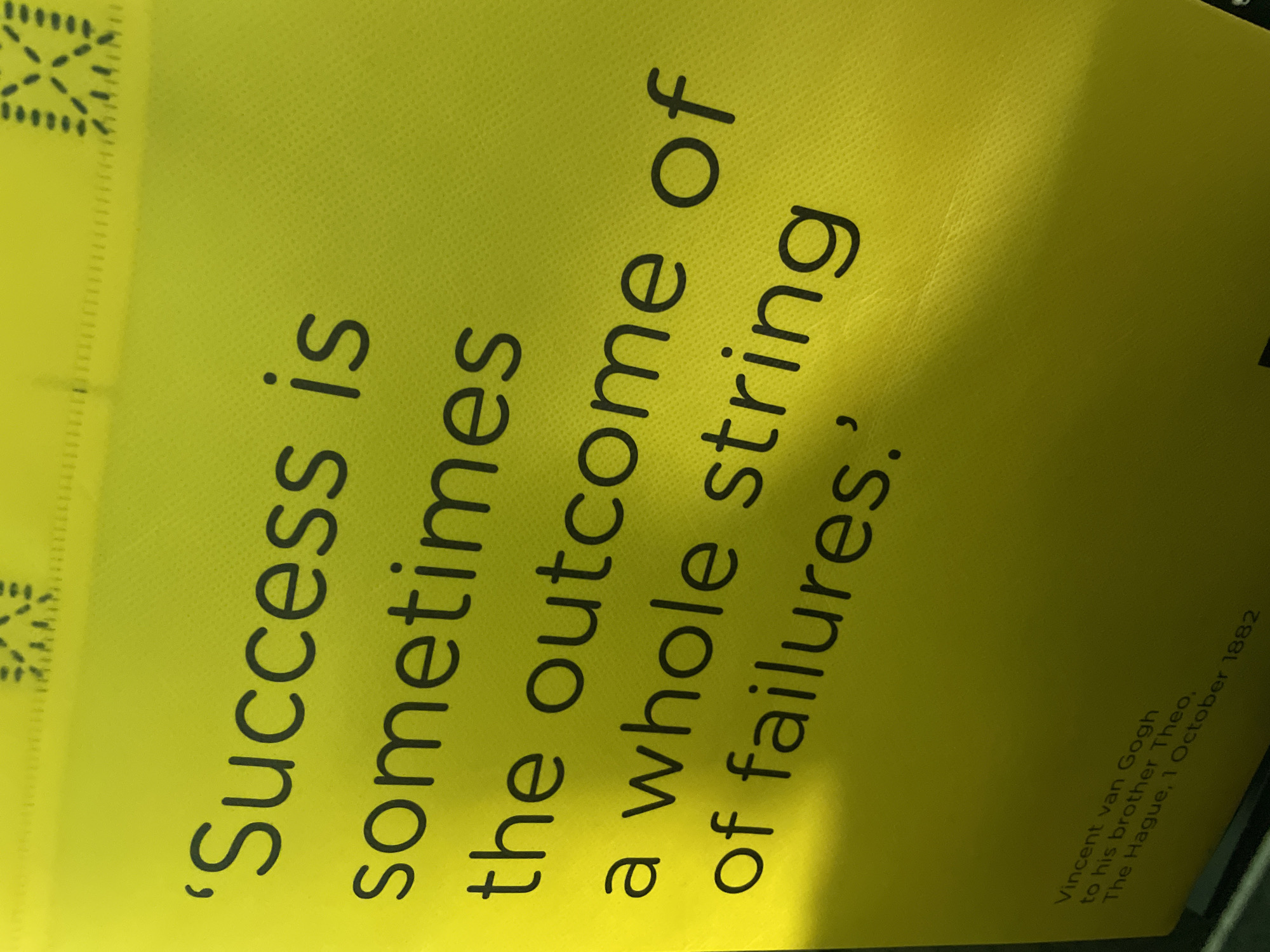 A close-up, eye-level, slightly angled medium shot shows a bright yellow surface with black text. The text reads: 'Success is sometimes the outcome of a whole string of failures.' Below this, in smaller font, it reads: Vincent van Gogh to his brother Theo, The Hague, 1 October 1882. The surface appears to be a fabric or textured paper, with a faint diagonal shadow cast across it from the left. There is a border of small, dark geometric patterns along the top edge of the frame and a hint of a dark object or surface at the very bottom. No people are visible in the frame.