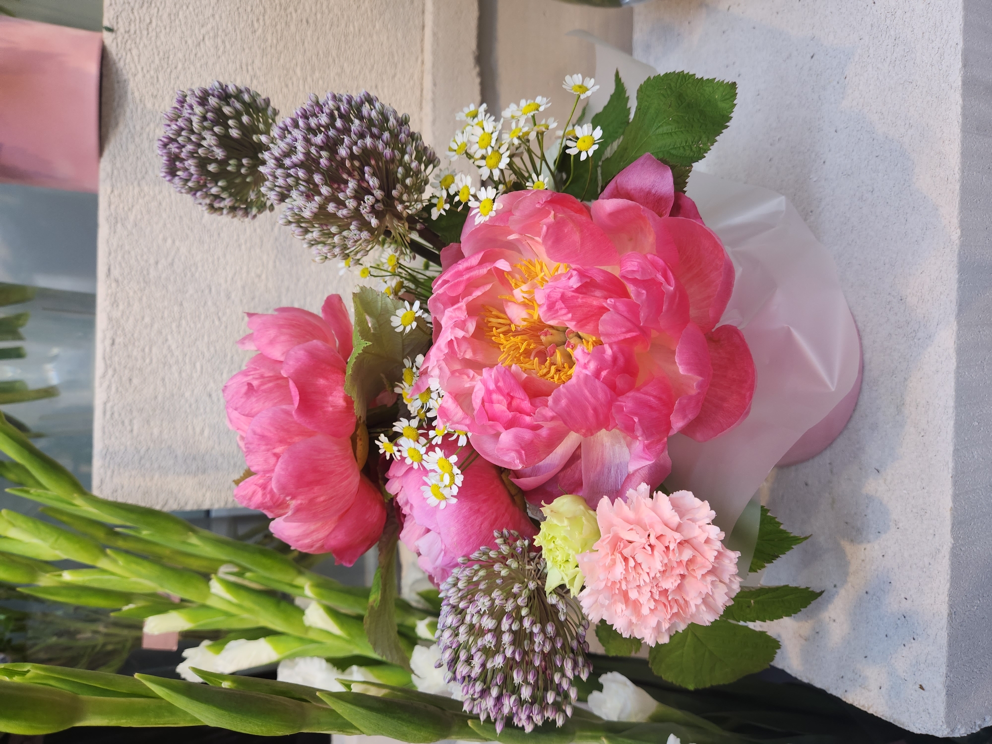 A bouquet of pink peonies, white carnations, and small white flowers with yellow centers is displayed in a light pink vase. The bouquet also includes purple ornamental onion flowers and green gladiolus stalks. The arrangement is placed on a textured grey surface, likely a countertop or display stand, with a light-colored textured wall in the background. The scene appears to be indoors, possibly in a flower shop or a home setting in Moscow, Russia.