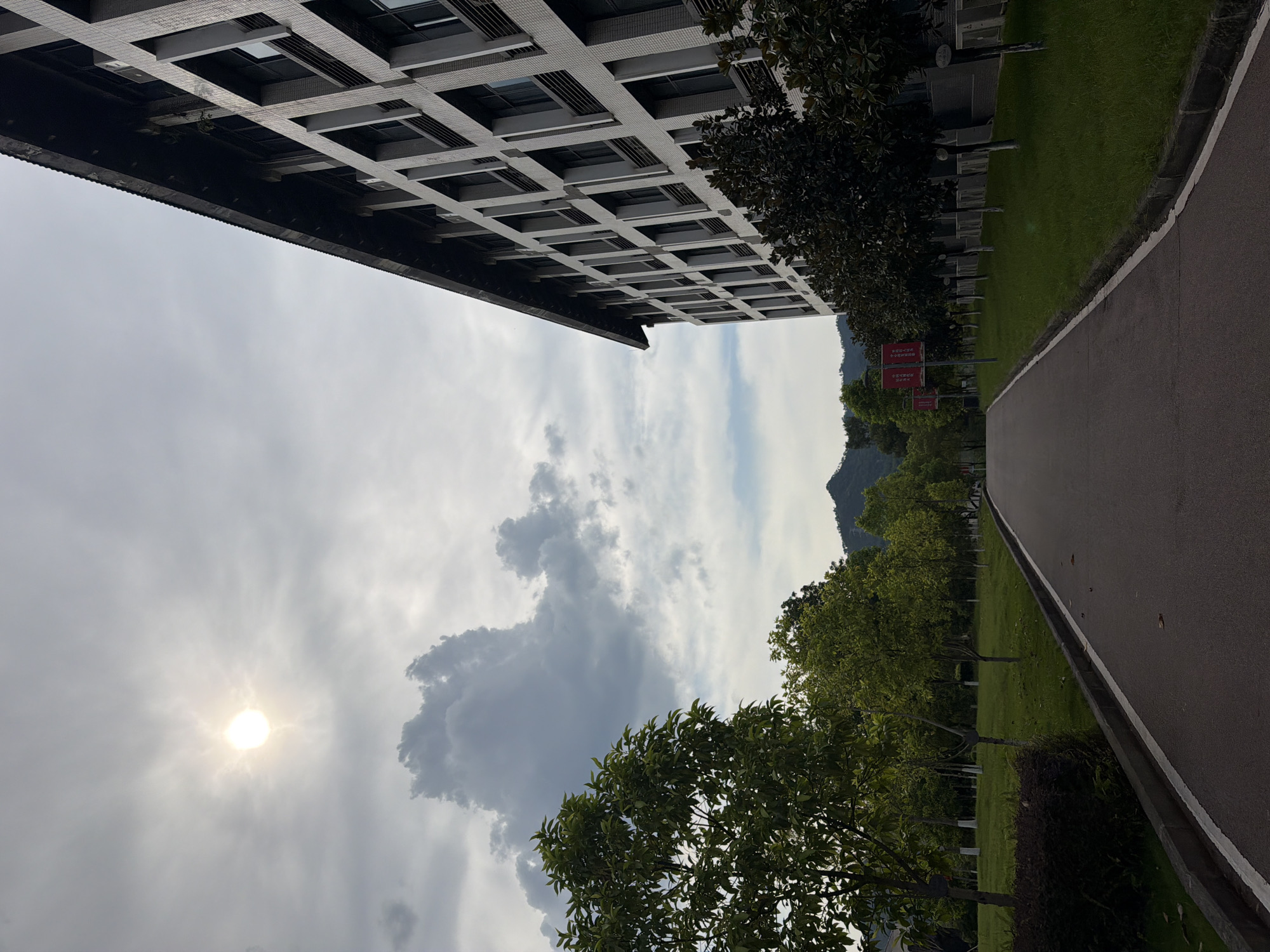 The image depicts a pathway leading through a green campus environment with a modern building on the right side. The sun is partially obscured by clouds in the upper left quadrant, casting a diffused light. On the left, lush green trees and manicured lawns are visible, creating a serene atmosphere. The building on the right is a multi-story structure with a grid-like facade of windows and balconies, suggesting an institutional or office setting. The pathway is paved and bordered by grass and trees, with some red banners hanging from poles further down the path. The overall scene is one of a well-maintained urban campus, possibly located in the city of Jiangjin, China, given the context provided. There are no people visible in the image, and no specific actions or interactions are depicted. The environment appears to be one of natural beauty integrated with modern architecture.
