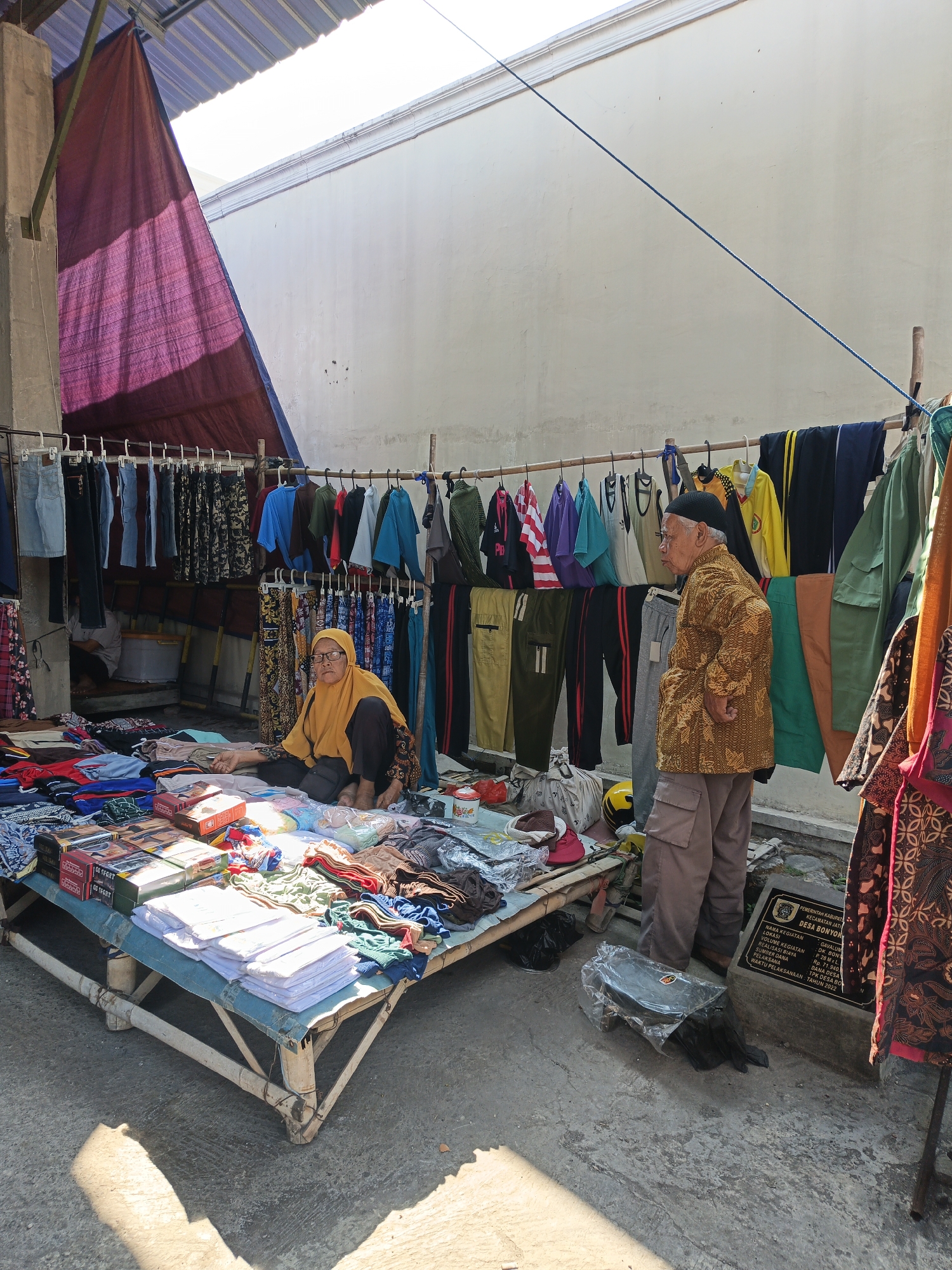 In Jatinom, Indonesia, a marketplace scene unfolds under a canopy with a red tarp. Two individuals are present: a woman in a yellow headscarf and dark clothing is seated on the ground amidst a spread of clothing and boxed items. Beside her, an elderly man, wearing a patterned batik jacket and brown pants, stands by a rack of hanging clothes. The market stall features a raised platform covered in a blue tarp, displaying various garments and boxes. The background consists of a light-colored wall and a metal roof structure. The ground is concrete, with shadows cast by the sunlight. A stone plaque with Indonesian text is visible near the man.