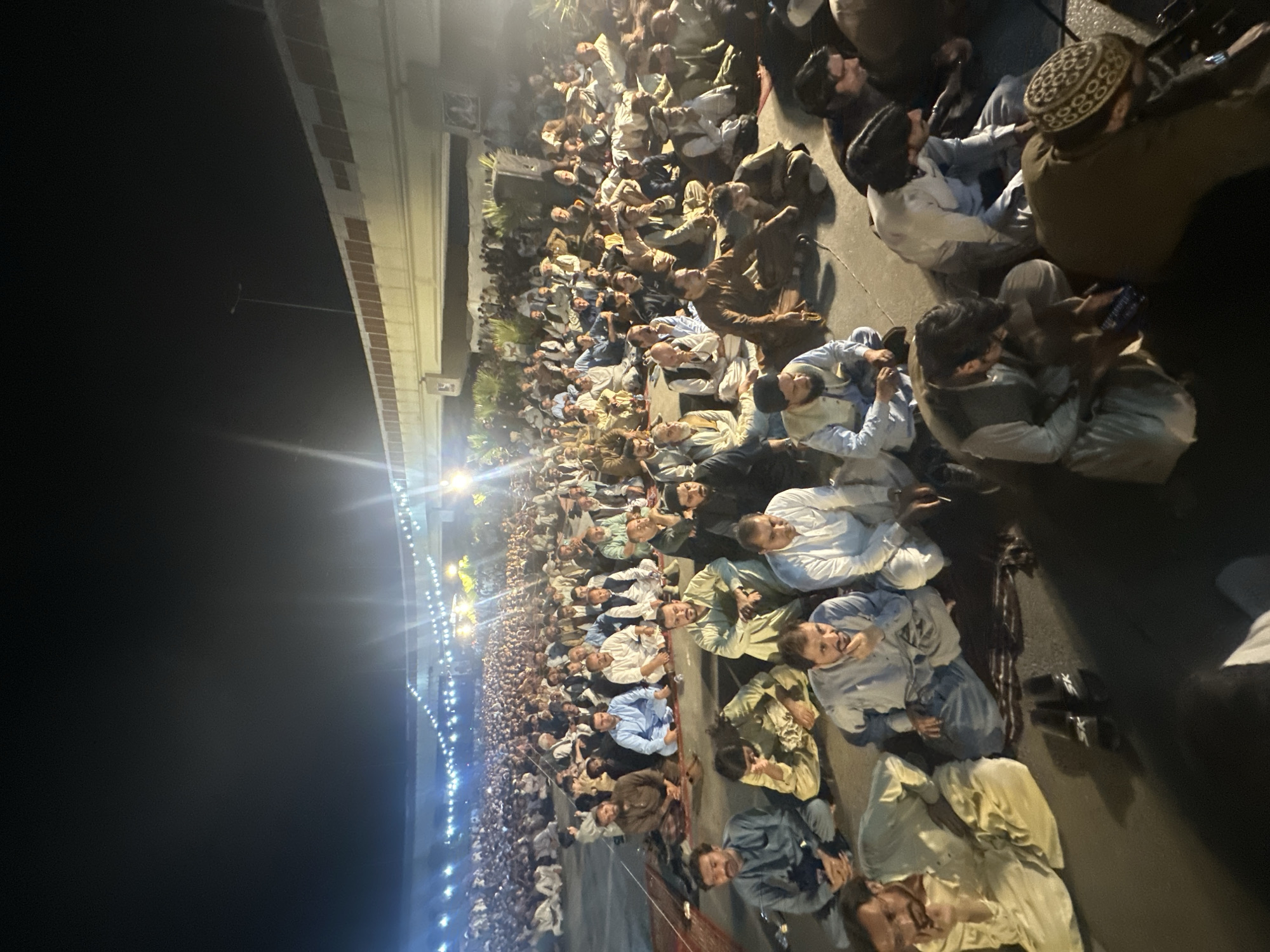 An aerial shot captures a large gathering of men seated on the ground, facing forward, outdoors at night. The scene is illuminated by strings of lights suspended overhead and light from a highway overpass. The individuals are dressed in traditional Pakistani attire, predominantly shalwar kameez in various neutral colors. They are positioned on carpets and directly on the ground, indicating a communal event or prayer. The atmosphere appears solemn, with many individuals in contemplative poses. The location is identified as Quetta City Tehsil, Pakistan, suggesting this is a public gathering in that area. The background shows the underside of an overpass, with more lights and a dense crowd extending into the distance.