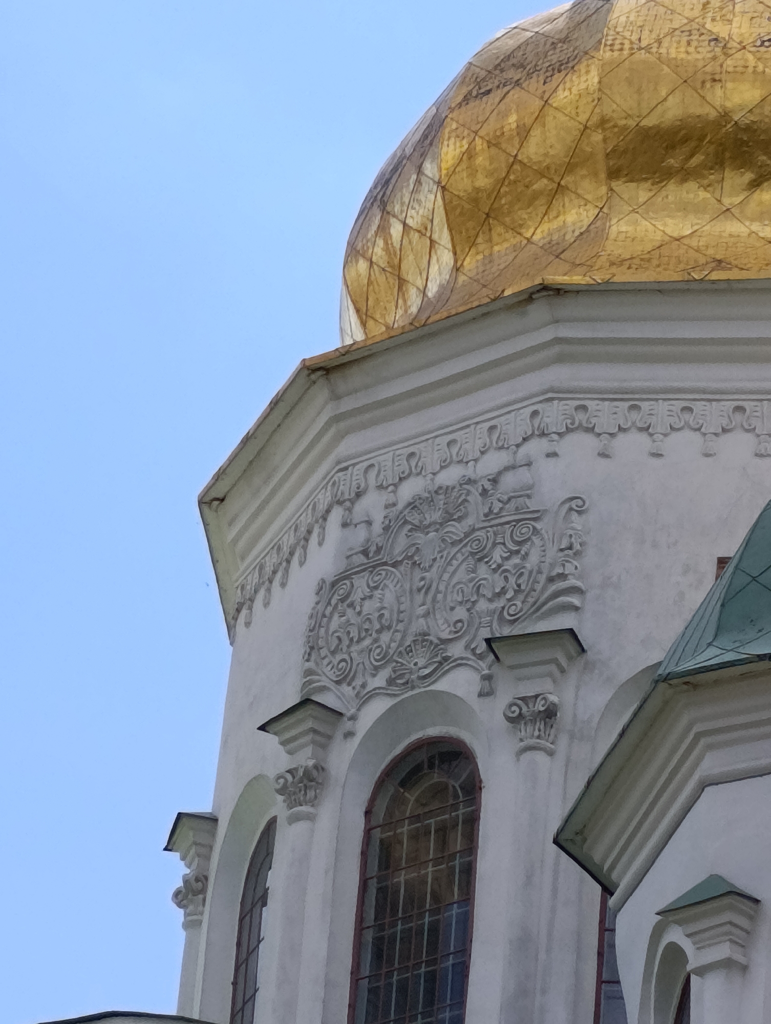 This media file is a close-up, low-angle shot of the upper section of a white building with a golden dome and a green roof. The building appears to be a religious structure, possibly a church or cathedral, given the prominent golden dome and architectural style. The dome is covered in a diamond-patterned material that reflects the light, giving it a shimmering appearance. Below the dome, the white facade of the building features elaborate decorative carvings. These carvings include intricate scrollwork and geometric patterns, indicative of classical or baroque architectural influences. There are also arched windows visible on the building's surface, with dark frames and what appear to be metal grates. Small, decorative columns with volute capitals are situated between the windows and also support projecting elements. A portion of a green, multi-tiered roof is visible on the right side of the image. The sky in the background is a clear, bright blue, suggesting it is daytime with fair weather. There are no people visible in the frame. The overall impression is one of grandeur and historical architecture.