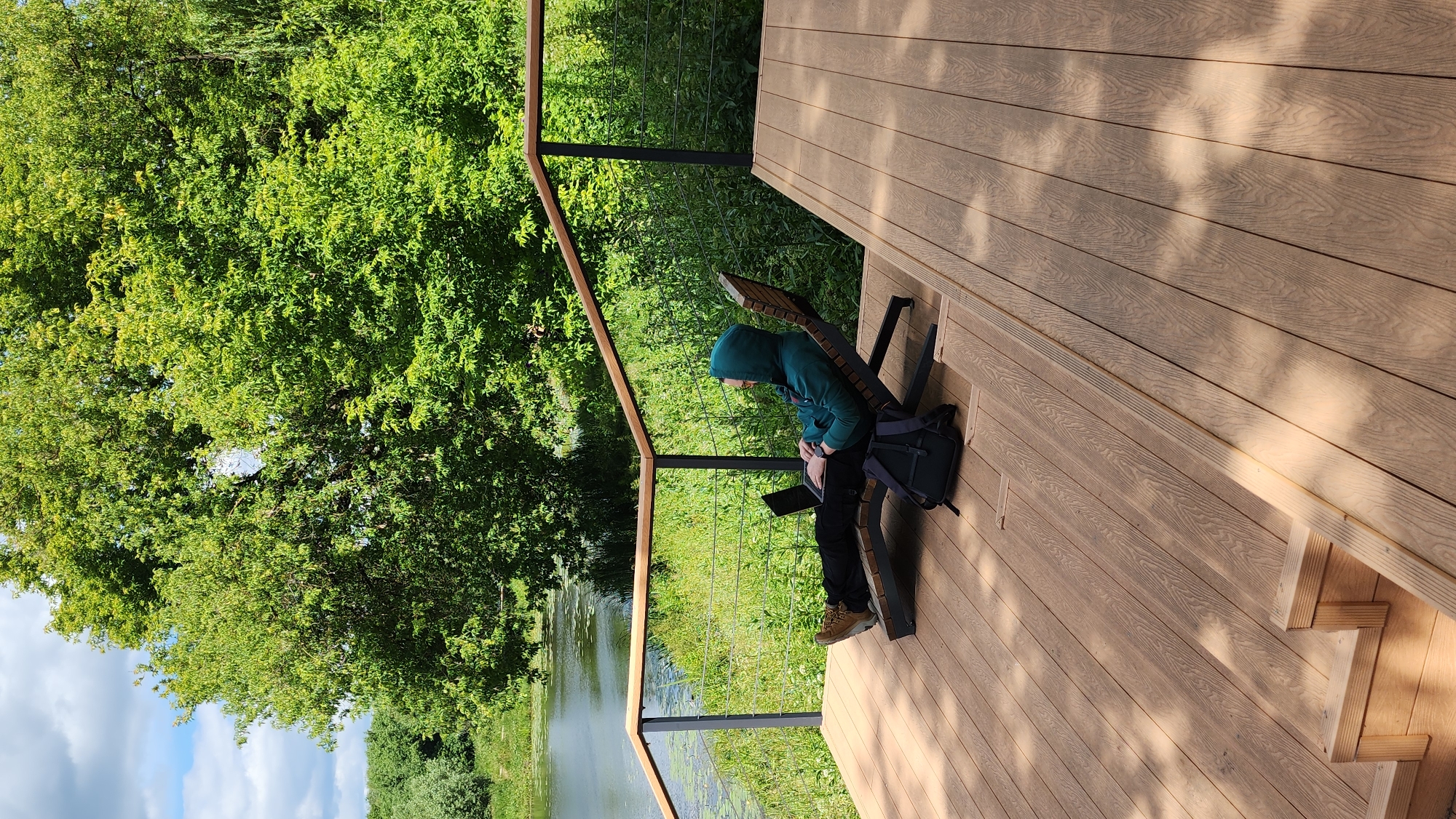 A person is sitting on a chaise lounge on a wooden deck, looking at a laptop. The person is wearing a green hooded sweatshirt and black pants. A black backpack is on the deck next to the chaise lounge. The deck overlooks a body of water surrounded by lush green trees. The sky is partly cloudy. The location is a park in Moscow, Russia.
