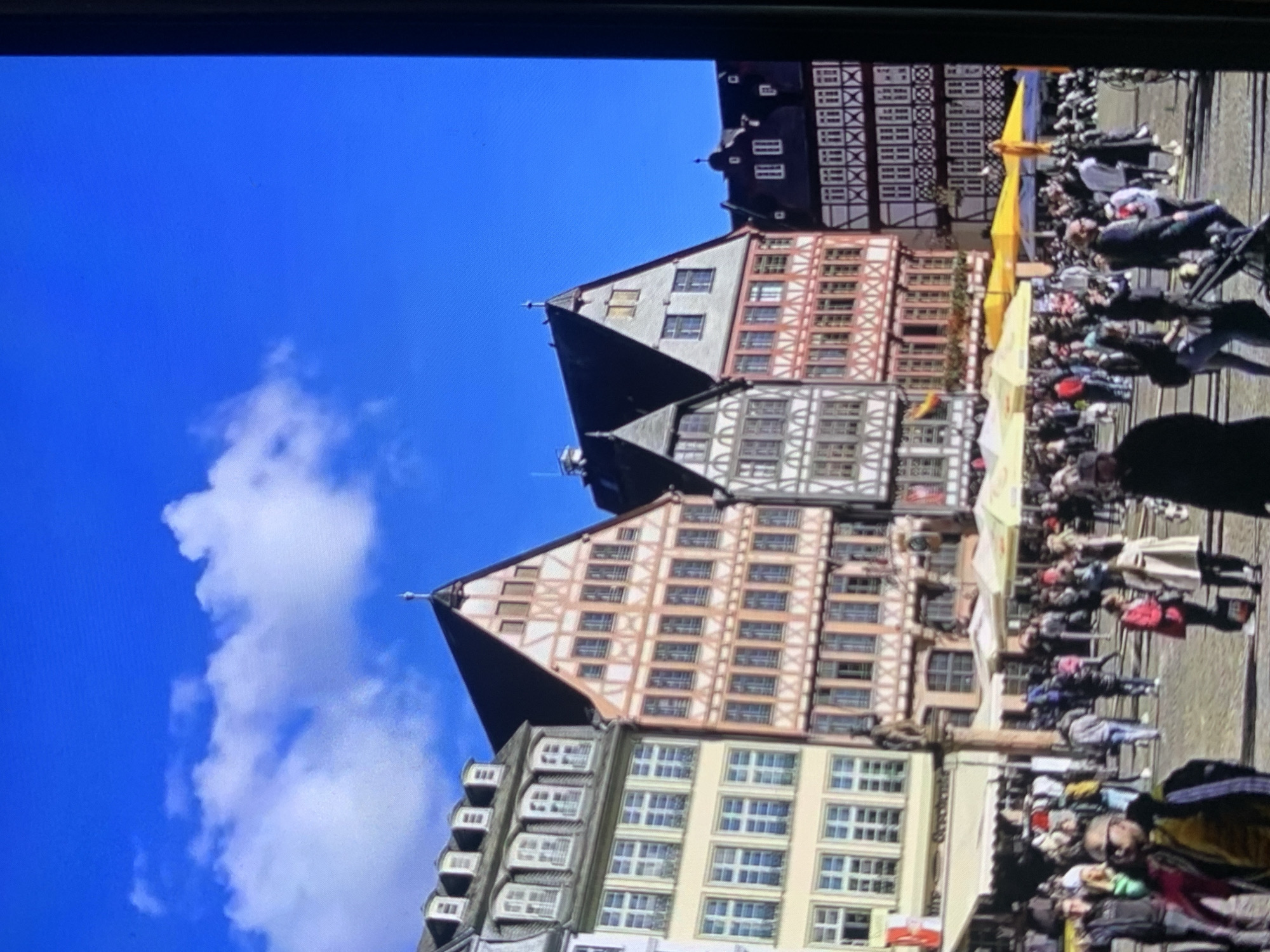 The image depicts a bustling town square filled with people. In the background, several traditional European buildings with timber framing and ornate facades are visible. The architecture suggests a historical European city. The sky is a vibrant blue with scattered white clouds. In the foreground, a crowd of people are walking and gathered, some under yellow umbrellas, indicating an outdoor market or gathering. There are also some trees and streetlights visible. The overall scene conveys a lively atmosphere in a picturesque urban setting. The location is identified as the city of Stavropol, Russia.