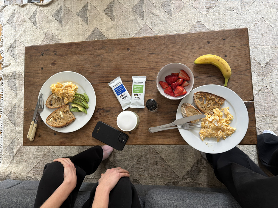The scene depicts a breakfast or brunch setup on a rustic wooden coffee table in an indoor residential setting in Paris, France.
On the dark brown wooden table, two individual meals are arranged. The upper white ceramic plate holds a portion of scrambled eggs, two slices of toasted artisan bread, and fanned slices of avocado. A fork and knife with light-colored handles are positioned above this plate. Next to this, a black smartphone in a protective case rests face down. Below the phone, a cylindrical light green container with a white lid is present. Two packaged "Hero" brand protein bars, one with a blue label and one with a green label, are placed beside the container. A black pepper grinder or salt shaker is located near the center of the table. The lower white ceramic plate contains a similar portion of scrambled eggs and two slices of toasted artisan bread, with a silver-colored fork and knife resting on it. To the right of the lower plate, a white bowl filled with sliced red strawberries is visible. A single yellow banana with brown speckles lies beside the lower plate.
Two individuals are partially visible. On the left, one person is seated on a grey upholstered sofa, wearing black pants and a light pink sock or slipper on their right foot. Their hands are clasped over their knees. Another person's legs, also clad in black pants with a white sock, are visible at the bottom left, positioned near the table.
The environment includes a light-colored rug with geometric triangular patterns in shades of beige, grey, and faint pink, covering the floor around the coffee table.