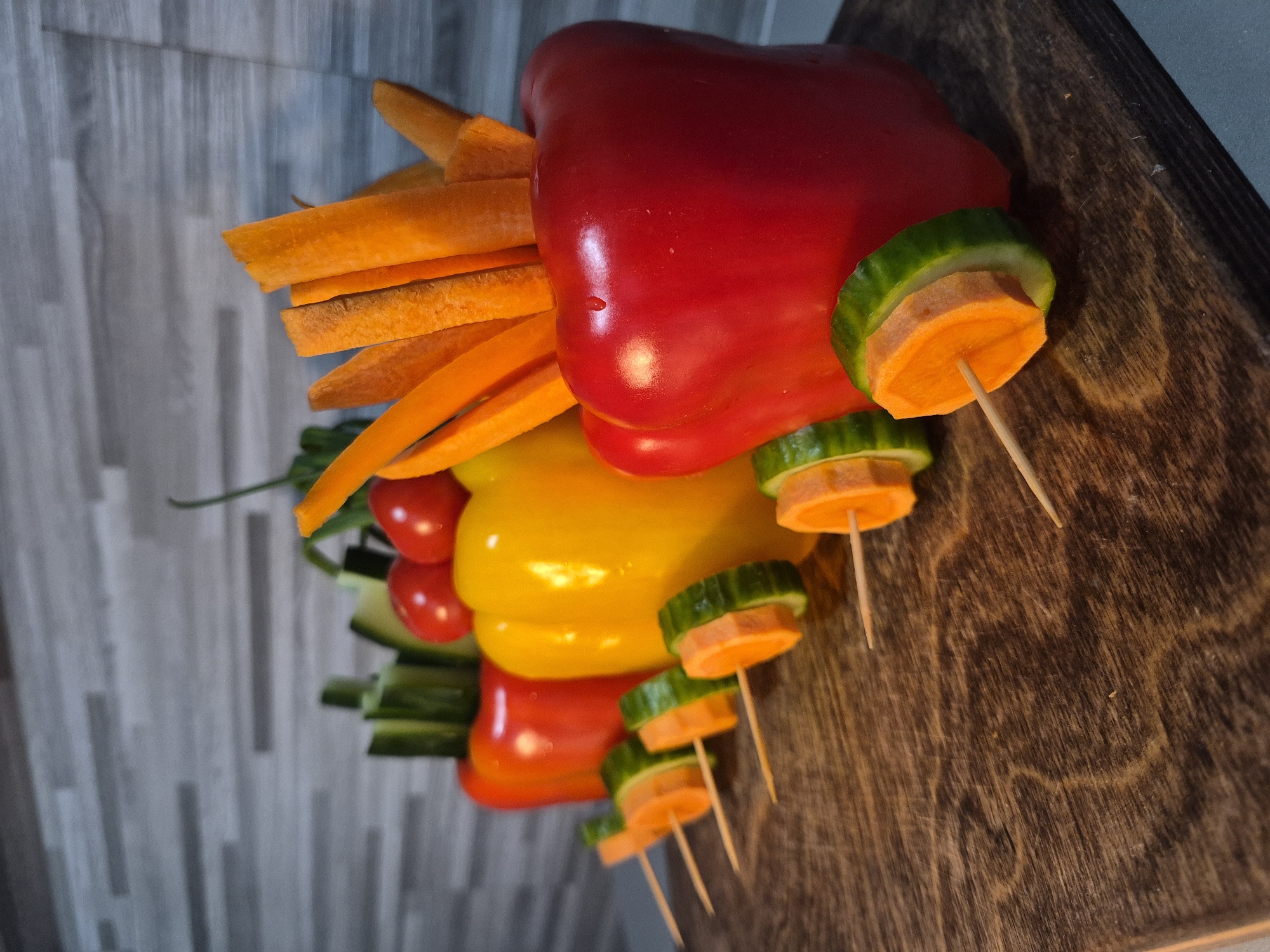 The image shows a colorful arrangement of vegetables resembling a car. The body is constructed from stacked red, yellow, and orange bell peppers. Carrots are cut into strips and used as a portion of the front of the vehicle. A row of small tomatoes and pieces of cucumber are stacked at the rear. The wheels are constructed from alternating layers of cucumber and carrot, held together by toothpicks. The vegetables are arranged on a dark wooden surface. A tiled wall serves as the background. The location is unknown.