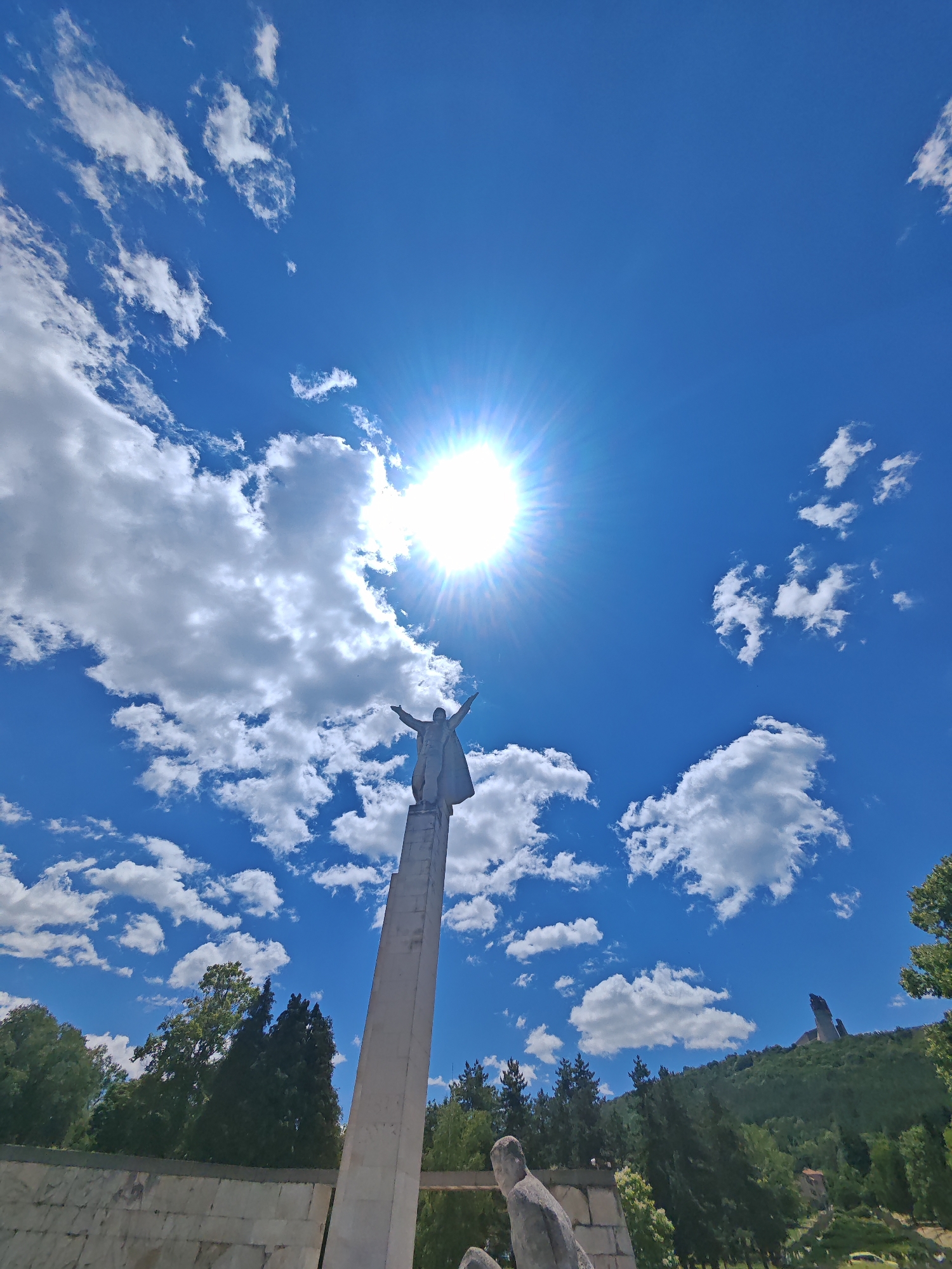 A low-angle, wide-shot photograph captures the Monument to the Founders of the Bulgarian State, situated on the Shumen Plateau in Shumen, Bulgaria. The monument, a monumental stone sculpture of a warrior with outstretched arms, stands atop a tall, slender column, silhouetted against a vibrant blue sky filled with scattered white clouds. The sun is prominently visible in the upper center of the frame, casting a bright glare and creating visible sunburst rays.
In the foreground, at the bottom of the frame, the edge of a stone wall and the base of another, partially obscured statue are visible. The midground is occupied by a dense forest of green trees, with a winding road or path partially visible in the lower right corner. In the background, on a distant hilltop, another dark, angular structure, possibly a memorial or a building, is discernible. The overall scene depicts a clear, sunny day with a dramatic sky above a significant historical monument.
