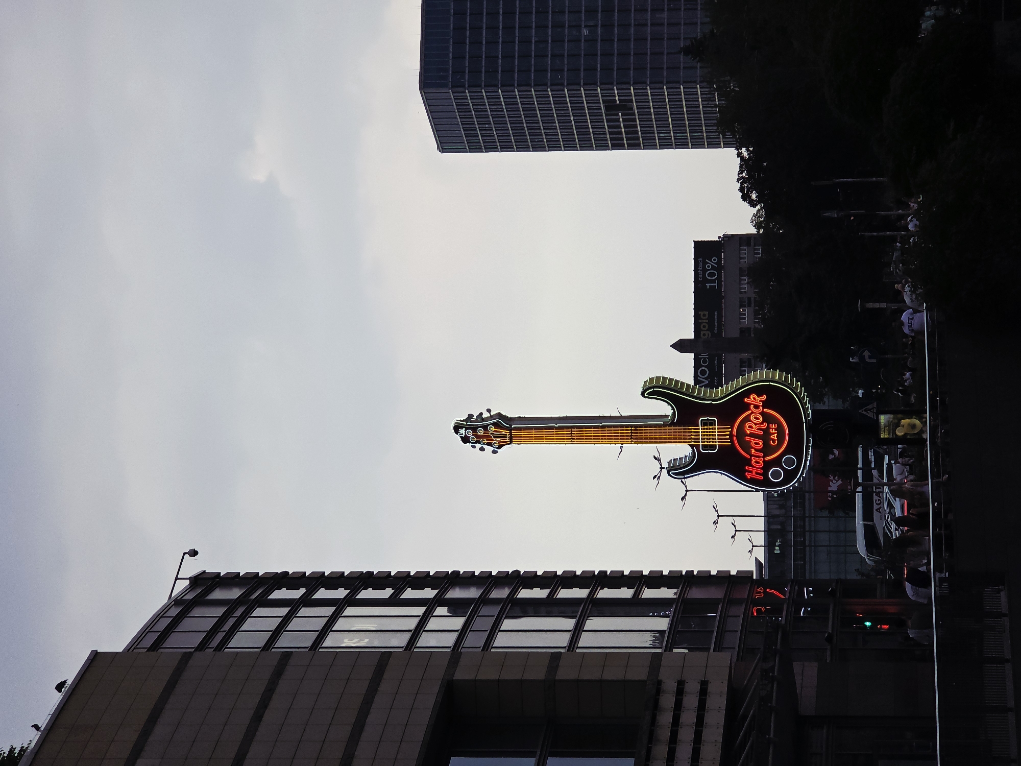 The image depicts an outdoor scene in Warsaw, Poland, featuring the Hard Rock Cafe sign prominently displayed as a large, illuminated guitar. The sign is positioned against a backdrop of urban architecture, including a modern building with a sloping, windowed roof, and a taller, grid-patterned skyscraper in the distance. People are present in the scene, near the base of the sign. A street is visible with traffic. A billboard is visible between the Hard Rock Cafe sign and skyscraper. The sky is overcast.