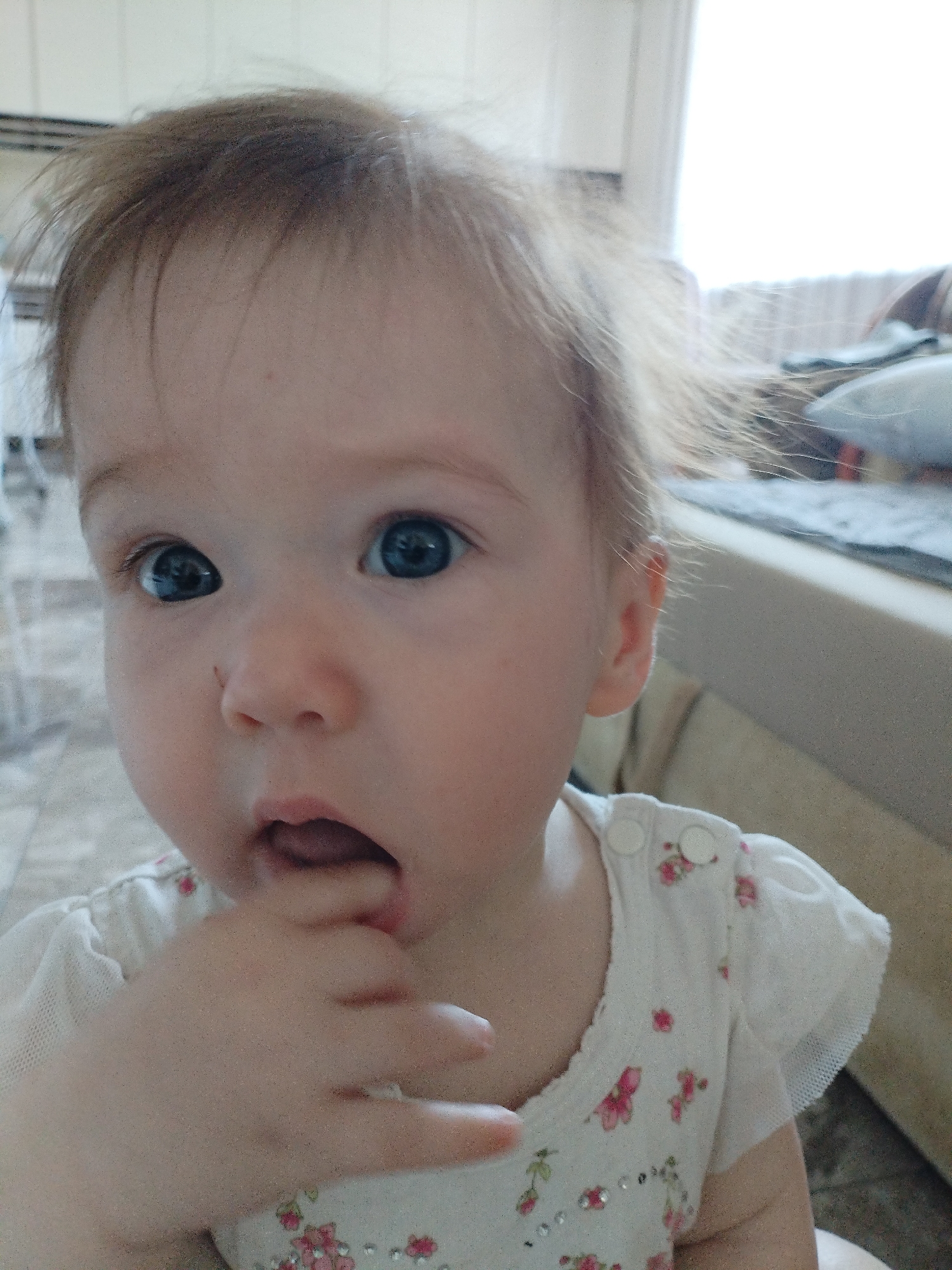 A frontal, waist-up portrait captures a light-haired infant or toddler with prominent blue eyes, positioned centrally within an indoor setting in the city of Vinogradny, Russia. The child's right hand, specifically the thumb and index finger, is partially inserted into their slightly open mouth. Their expression is attentive, with direct eye contact towards the viewer. The child is dressed in a white garment featuring small pink floral patterns and clear, reflective embellishments. The short sleeves exhibit a sheer, mesh-like fabric, and the shoulder strap is secured by two white buttons. The background is brightly lit, particularly on the right, suggesting the presence of a window with a light-colored sill and indistinct objects or textiles. The upper left background reveals faint horizontal lines against a light-toned surface.