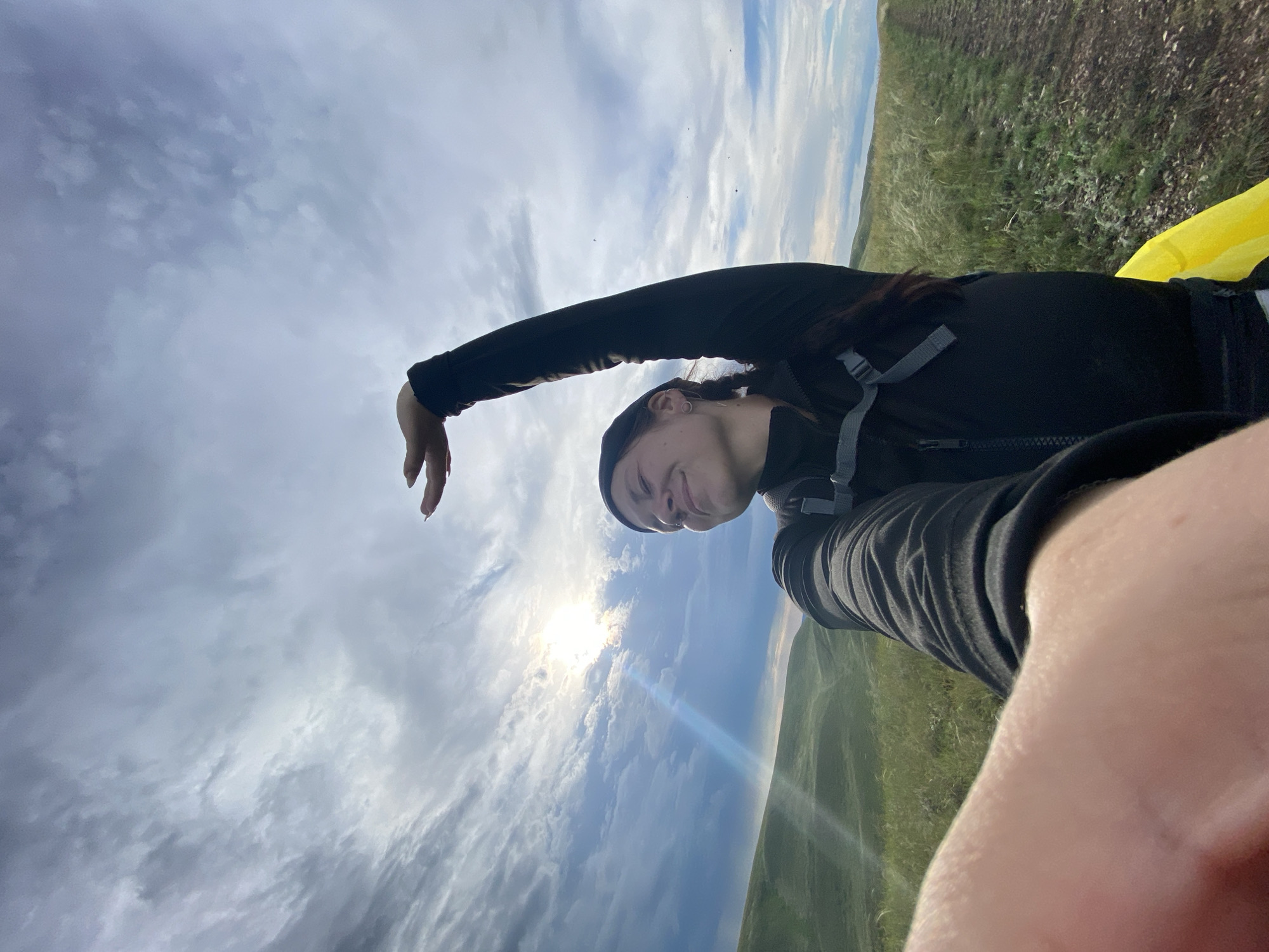 A woman with braided dark hair, wearing a black beanie, black long-sleeved shirt, and black pants, is captured in a selfie outdoors. She has her eyes closed and a slight smile, with her right arm raised above her head, palm open, as if in a graceful pose. She is wearing a backpack with grey straps. The background features a vast, cloudy sky with the sun peeking through, casting a lens flare and a subtle rainbow effect. Rolling green hills, covered in grass and wildflowers, stretch out behind her, and a dirt path is visible to her right. The scene suggests an outdoor adventure or hike in a natural landscape.