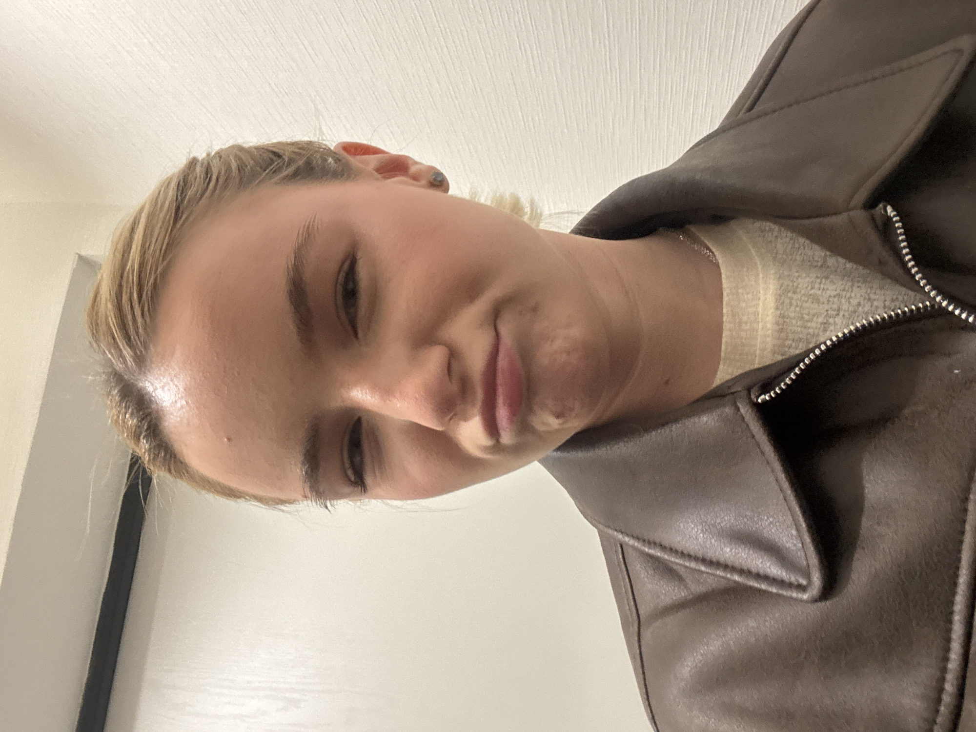 A blonde-haired person wearing a brown jacket is in an indoor environment. The person is looking at the camera with a slight frown. The background appears to be a white wall. The location is Unknown, Unknown.