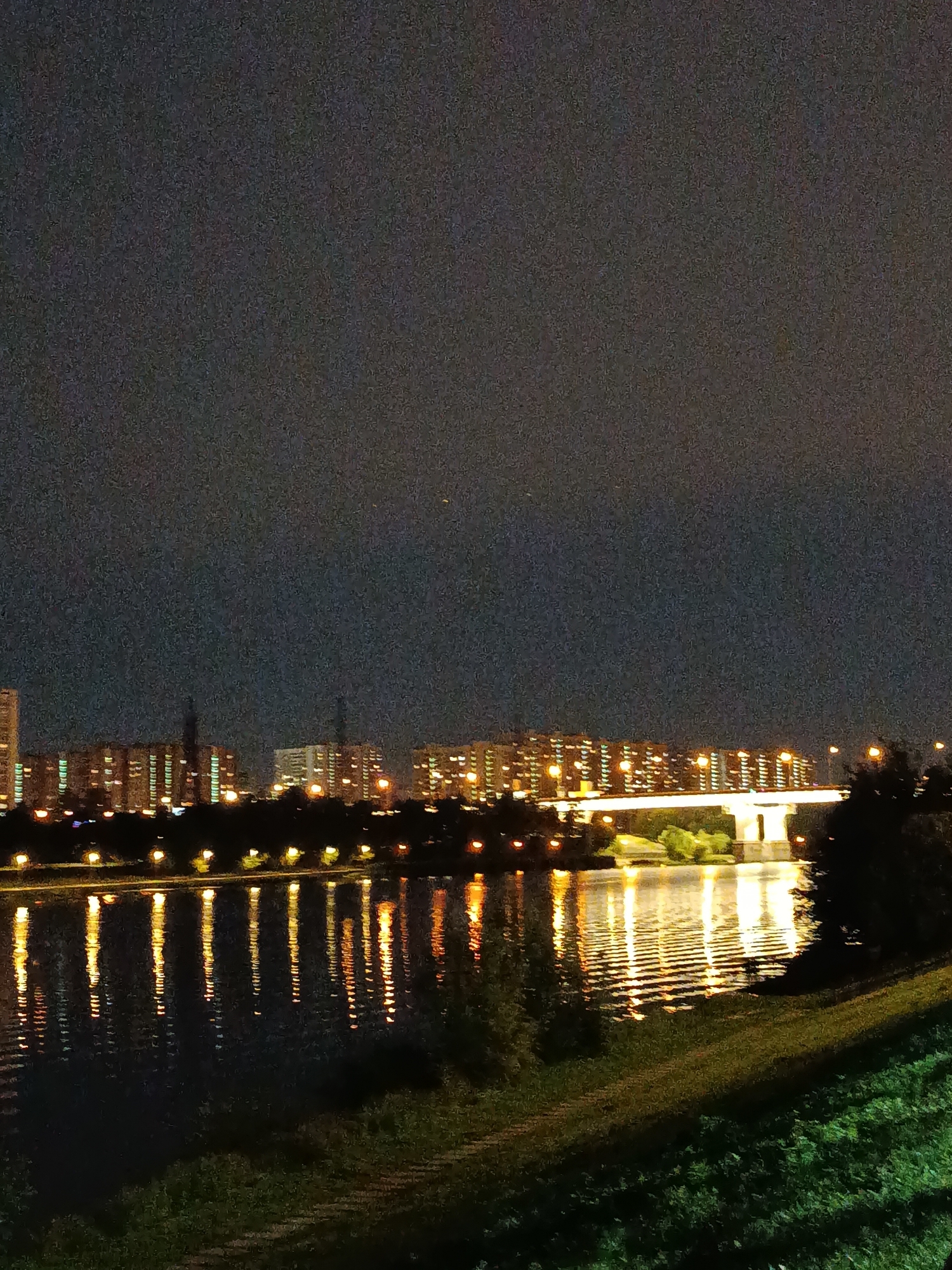 A nighttime, wide-angle, outdoor photograph depicts a city skyline across a body of water. The sky is dark with minimal visible stars, suggesting a heavily light-polluted environment. A line of residential buildings with illuminated windows stretches across the middle ground. A bridge with illuminated supports spans the water in front of the buildings. The water reflects the lights from the buildings and bridge, creating elongated streaks of light on its surface. The foreground shows a grassy embankment sloping down towards the water. The overall impression is a view of an urban area at night.