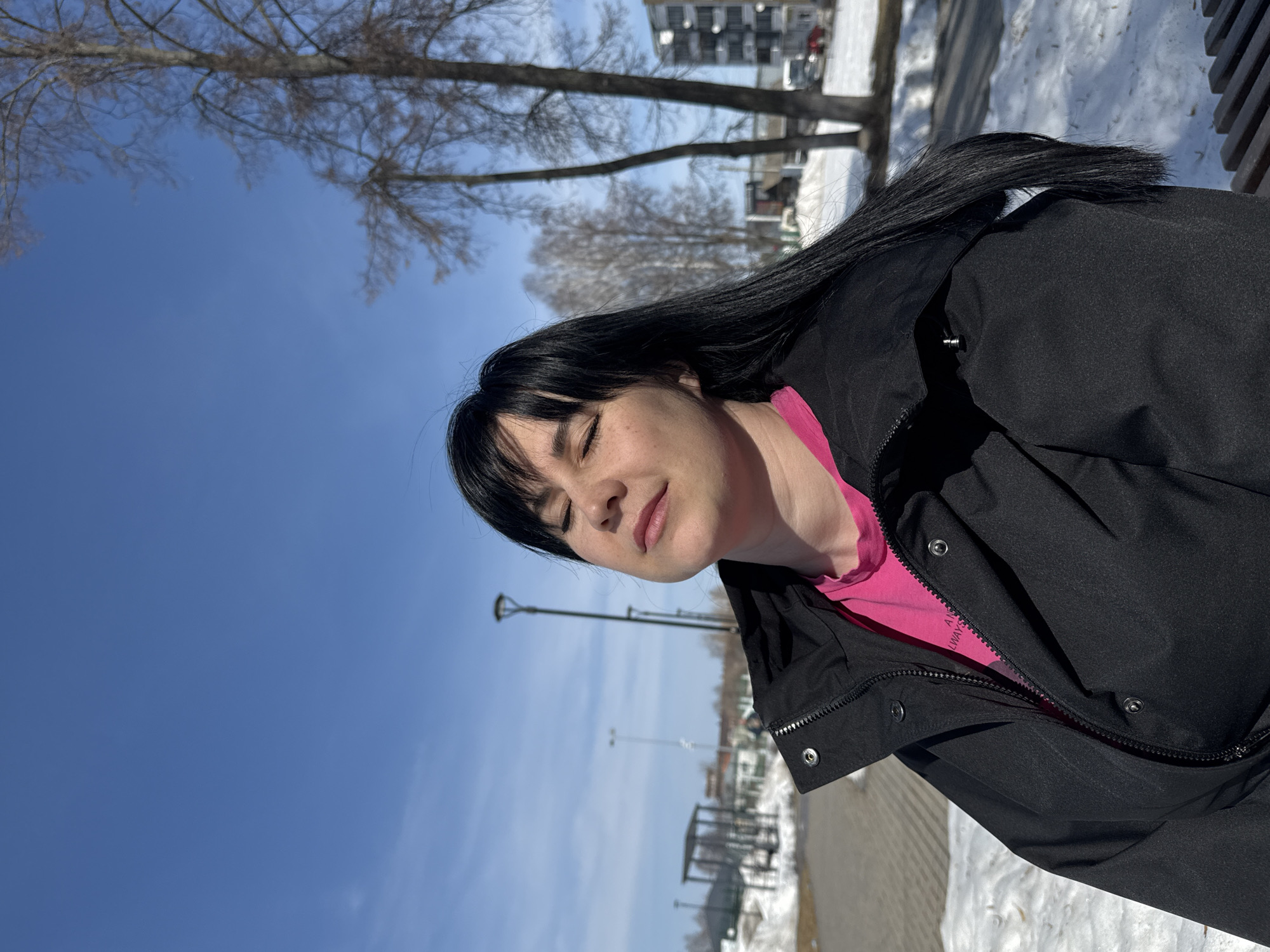 A young woman with black hair is sitting outside on a sunny day. She is wearing a pink shirt under a black jacket. The background includes bare trees, a blue sky, snow on the ground, benches, a street lamp, and a building. The image appears to have been taken in a park or public area in the city of Zav'yalovo, Russia.