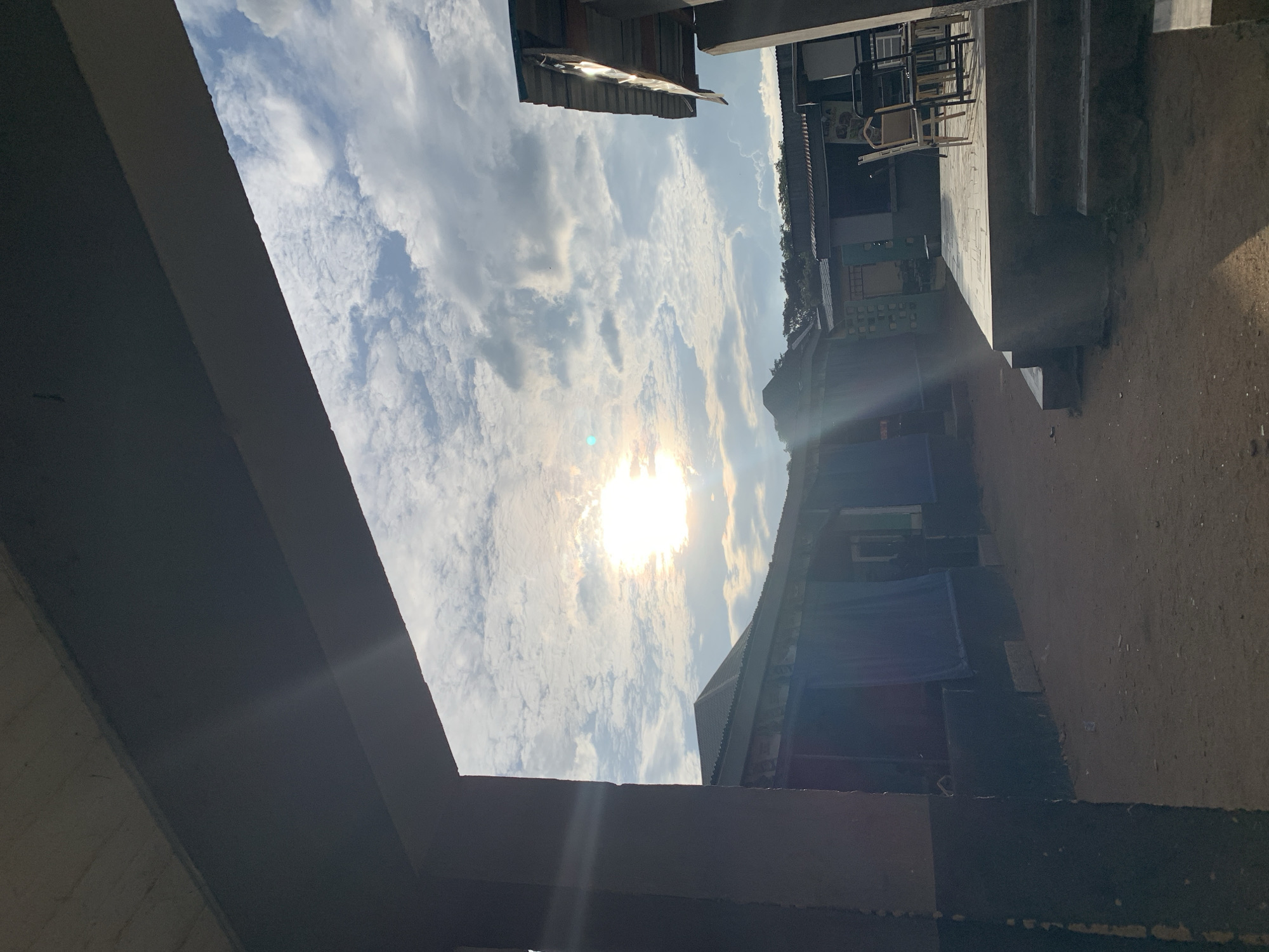 The image, captured in New Stadium, Nigeria, displays an outdoor scene framed by shadowed architectural elements. A wide, dark, flat concrete beam extends across the top of the frame, with a thick, dark, vertical concrete pillar on the left, both casting deep shadows. The ground in the midground is composed of dry, packed earth or dirt.
In the midground, a series of single-story buildings are arranged. On the left, a row of structures features dark-colored lower walls and dark fabric coverings over their window or doorway openings, topped with corrugated metal roofs. On the right, a raised concrete platform is accessible by three steps. Several chairs, including one light-colored, possibly beige chair and multiple dark-framed chairs (some stacked), are positioned on this platform. Behind the platform, another building with a corrugated metal roof displays a partially visible sign, possibly reading "Lady A."
The background reveals a sky filled with scattered white clouds, primarily cumulus and cirrocumulus formations. The sun, a brilliant white circular light source, is prominently located in the upper-left portion of the sky, emitting significant lens flare. A line of distant green foliage or trees is visible on the horizon, above the roofs of the buildings. No individuals are present in the scene. The strong backlighting from the sun creates stark contrasts between the brightly lit sky and the shadowed foreground and midground structures.