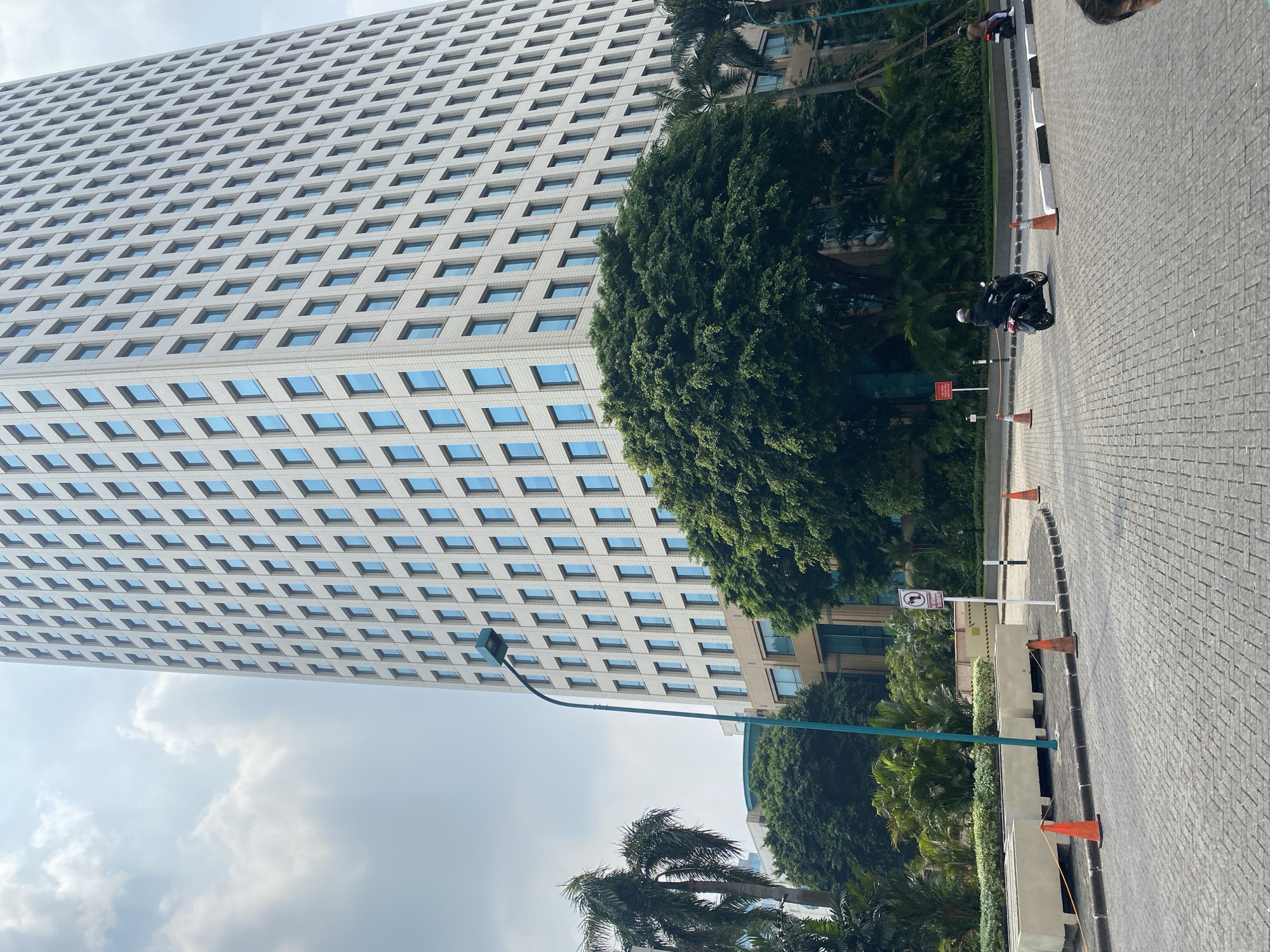 This is a daytime, low-angle outdoor shot of a modern, white-tiled skyscraper with numerous blue-tinted windows. The building dominates the background. In the mid-ground, a large, dense green tree stands in front of the skyscraper. To the left of the tree, a tall palm tree is visible. A streetlamp with a green pole is positioned in front of the tree and skyscraper. The foreground shows a paved road with a cobblestone-like pattern. Several orange traffic cones are placed along the edge of the road. Two people on motorcycles are visible on the road. One motorcycle is closer to the viewer and is black, ridden by a person wearing dark clothing and a helmet. Another motorcycle is further away and is lighter in color, with a rider in lighter clothing. The sky is partly cloudy. Based on the architectural style of the skyscraper and the presence of lush tropical vegetation, the location is consistent with Central Jakarta, Indonesia.