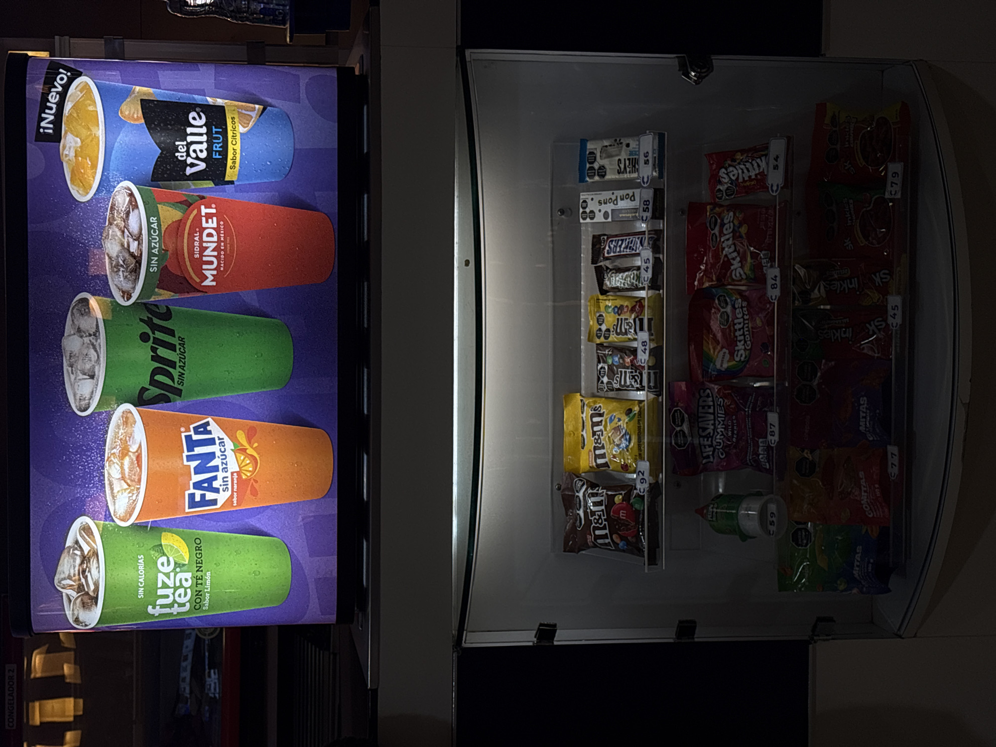 The image shows a vending machine in Puebla, Mexico. The vending machine has two sections, one for drinks and one for snacks. The drink section features advertisements for various beverages, including Del Valle Fruit, Mundet, Sprite, Fanta, and Fuze Tea. The snack section contains a variety of candies and snacks, including M&Ms, Snickers, Skittles, Inkles, and Ritas. Each item in the snack section has a price tag. The environment is dark, with the vending machine being the primary source of light. The overall scene appears to be taken indoors, possibly in a shopping mall or transportation hub.
