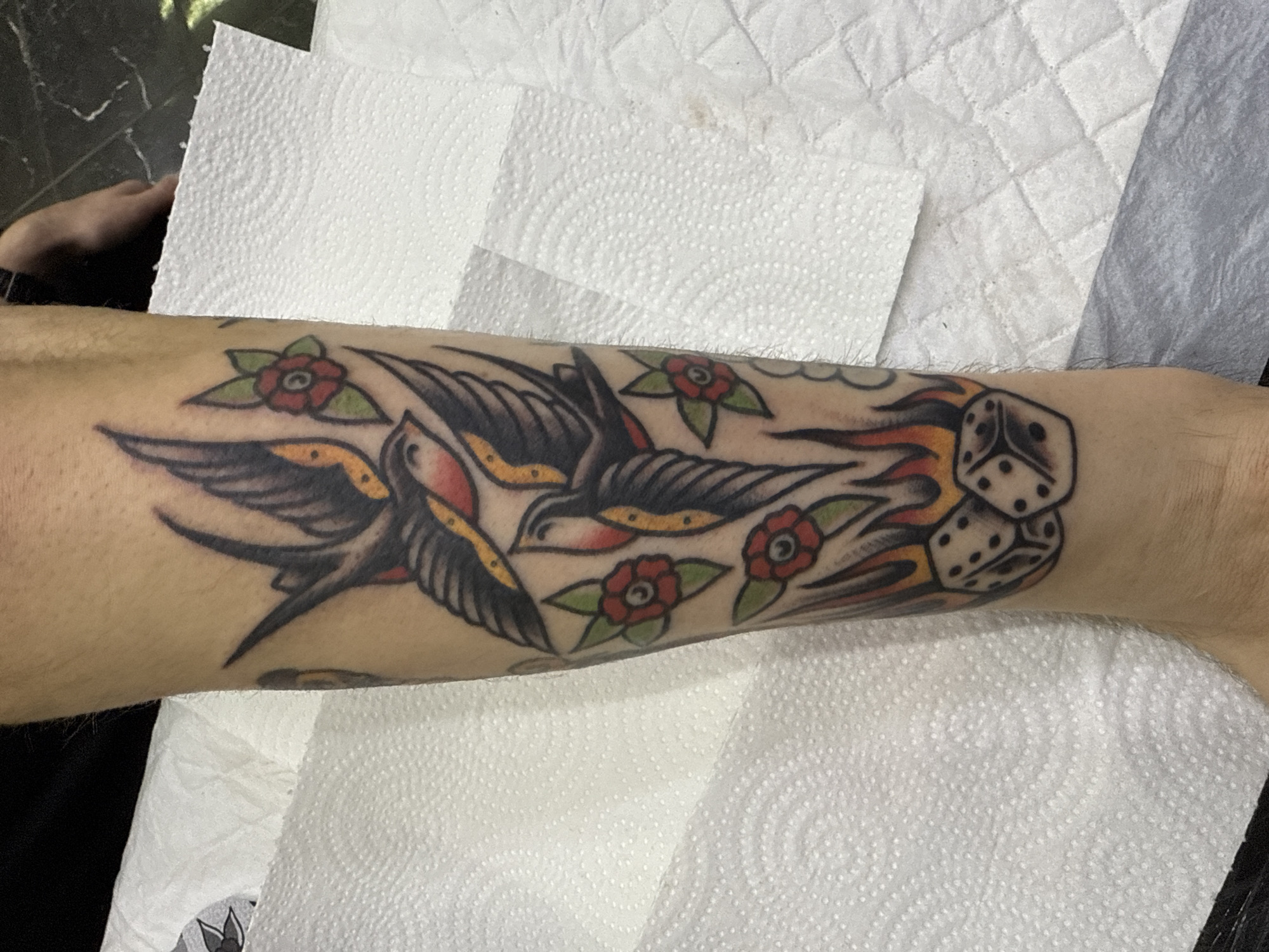 The image displays a close-up of a person's forearm adorned with a vibrant tattoo in a traditional American style. The central elements of the tattoo are two swallows, depicted with black bodies, yellow chests, and red accents on their throats and wings, shown in flight. They are surrounded by four red roses with distinct green leaves. To the right of the birds, a dynamic flow of red and orange flames transitions into a pair of black-and-white dice. One visible die face clearly shows five dots, while another face shows two dots.
The skin immediately surrounding the tattoo appears slightly reddened, indicative of a recently completed or in-progress tattooing session. The arm rests against or is positioned near several sheets of white, textured paper towels, commonly used in tattooing environments. A glimpse of a dark surface, possibly clothing or a table, and a partial view of another person's hand are visible in the upper left corner of some crops. The lighting is bright and consistent, suggesting an indoor setting. There is no discernible text visible within the image.
