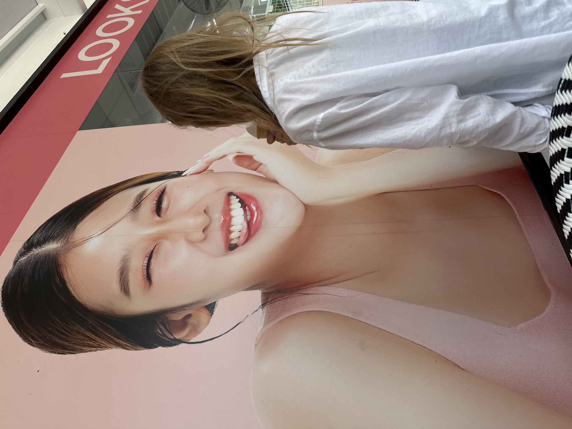A young woman with shoulder-length blonde hair is standing in front of a large mural advertising "LOOKS" on Koh Phangan, Thailand. She is wearing a loose-fitting white long-sleeved shirt and is holding a pale cone-shaped object, possibly an ice cream cone, near her face. The mural depicts a close-up of a young Asian woman with dark hair pulled back, wearing a pink tank top. She has her eyes closed and is smiling, with her tongue sticking out playfully. The background of the mural is a soft pink. The scene appears to be outdoors, possibly in a shopping area, given the signage.