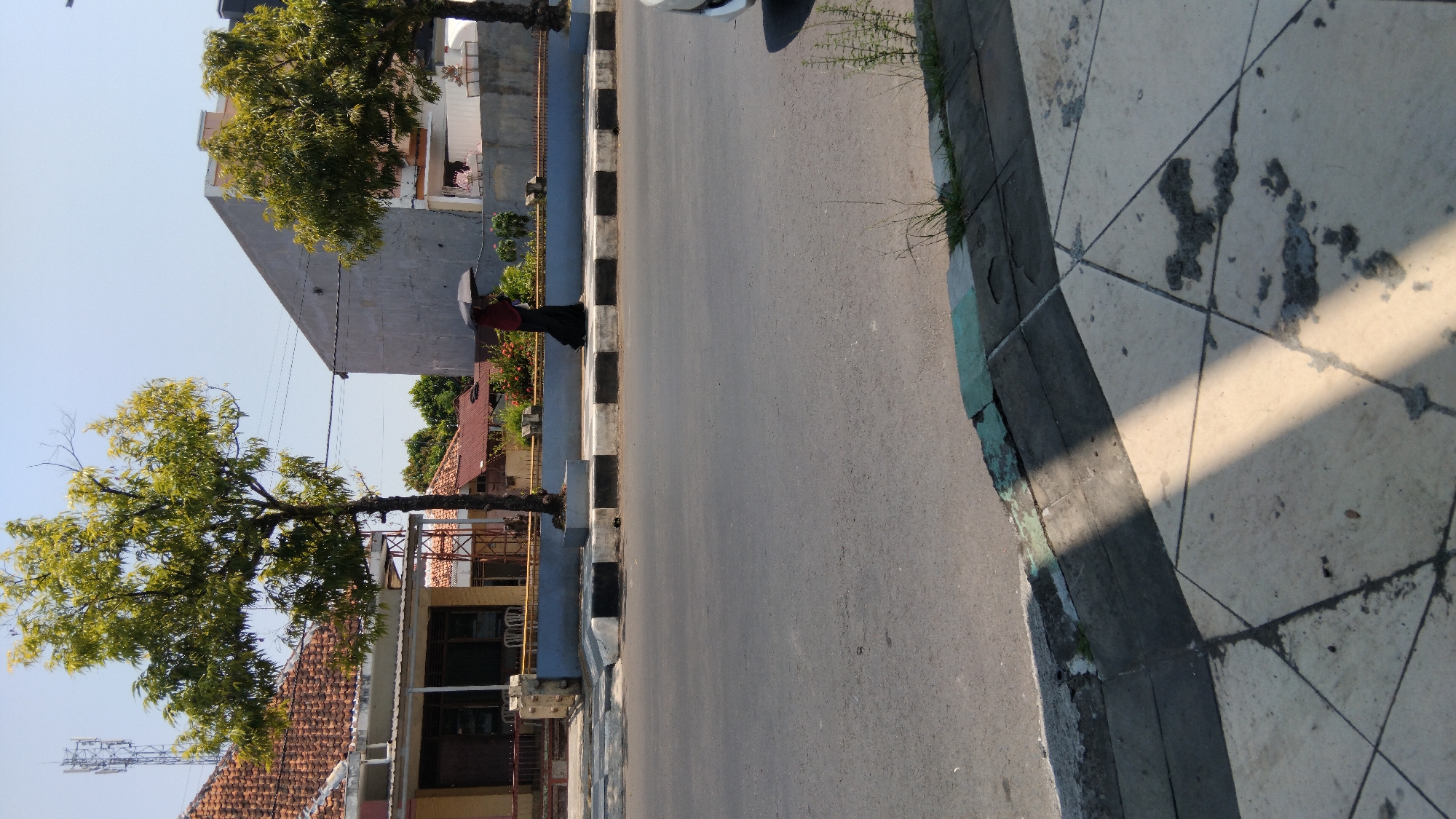 In Batang, Indonesia, a person walks along a sidewalk next to a road, holding a black umbrella. The person is wearing a dark abaya and a headscarf. The sidewalk is lined with a curb that has alternating black and white rectangular tiles. Beyond the curb is a paved road. On the other side of the sidewalk, there are trees, buildings with red tile roofs, and a grey building with a triangular facade. The sky is clear and blue. The sun is casting strong shadows on the sidewalk.